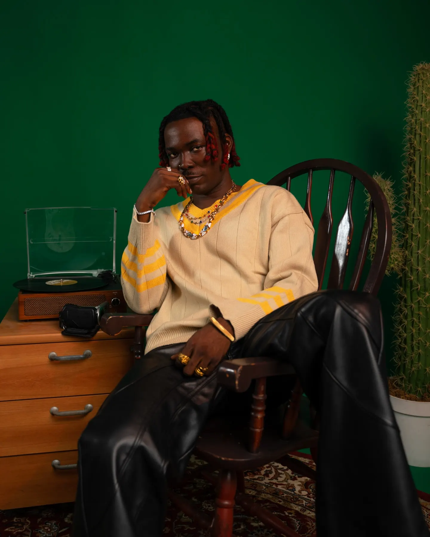 Musician in a cream V-neck sweater with yellow stripes and black leather trousers, sprawled in a wooden rocking chair beside a record player against a deep green wall.