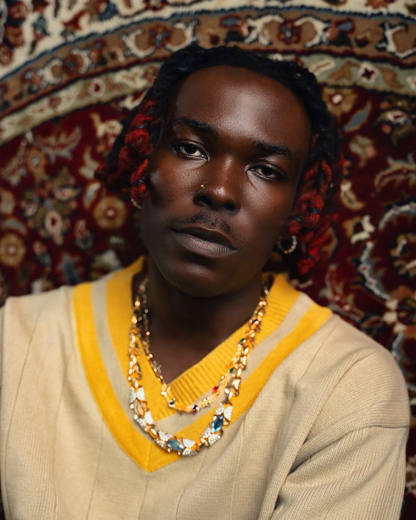 Close portrait of a musician with red-tipped braids in a cream-and-yellow cricket sweater layered with jeweled gold necklaces, gazing straight ahead before a red oriental rug.