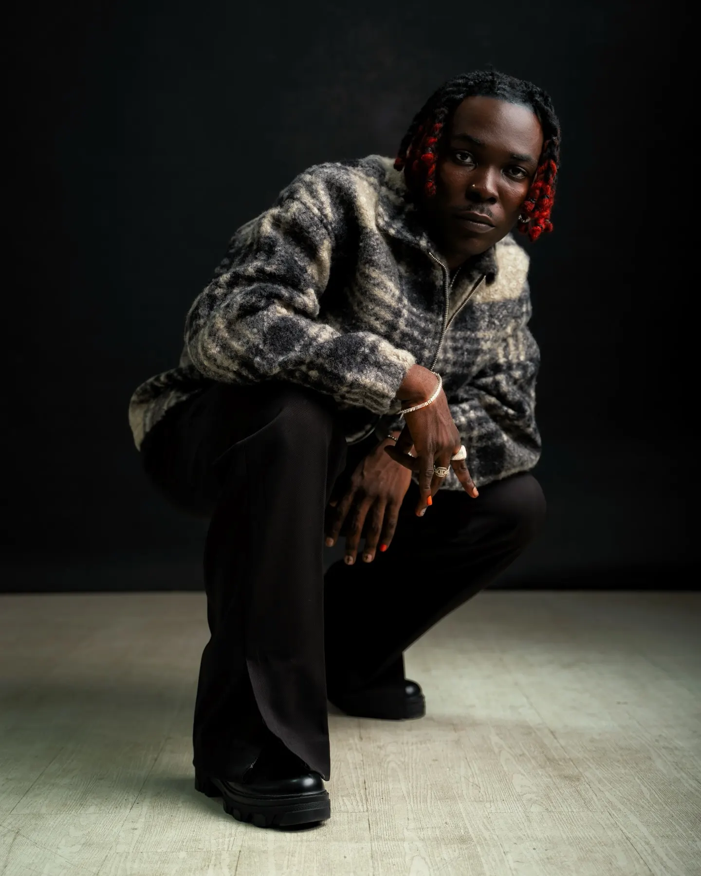 Musician in a grey plaid mohair jacket and wide black trousers crouching low on pale floorboards, fingers stacked with rings, against a black studio wall.