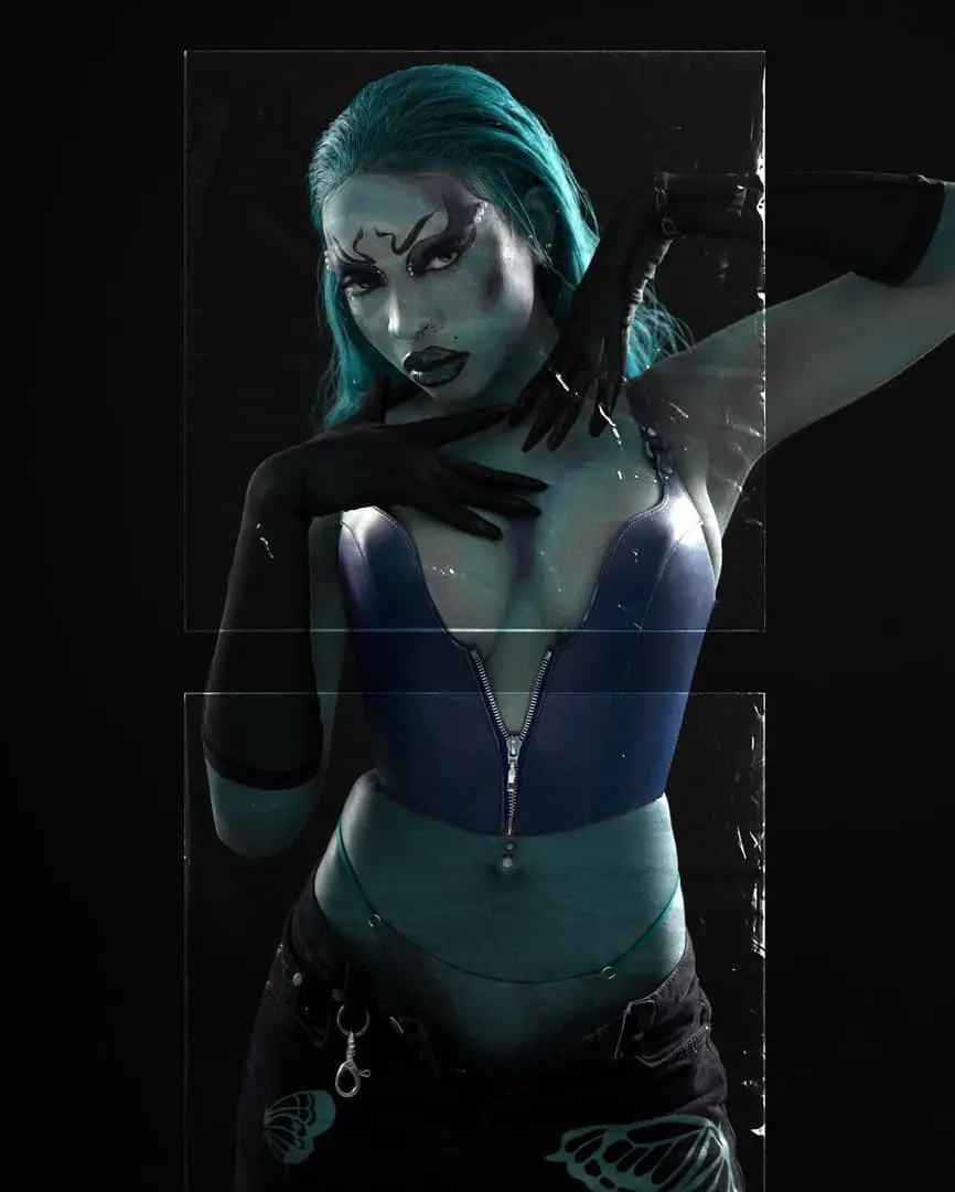 Model with teal hair, dramatic dark makeup and long black gloves poses in a zip-front corset against black.