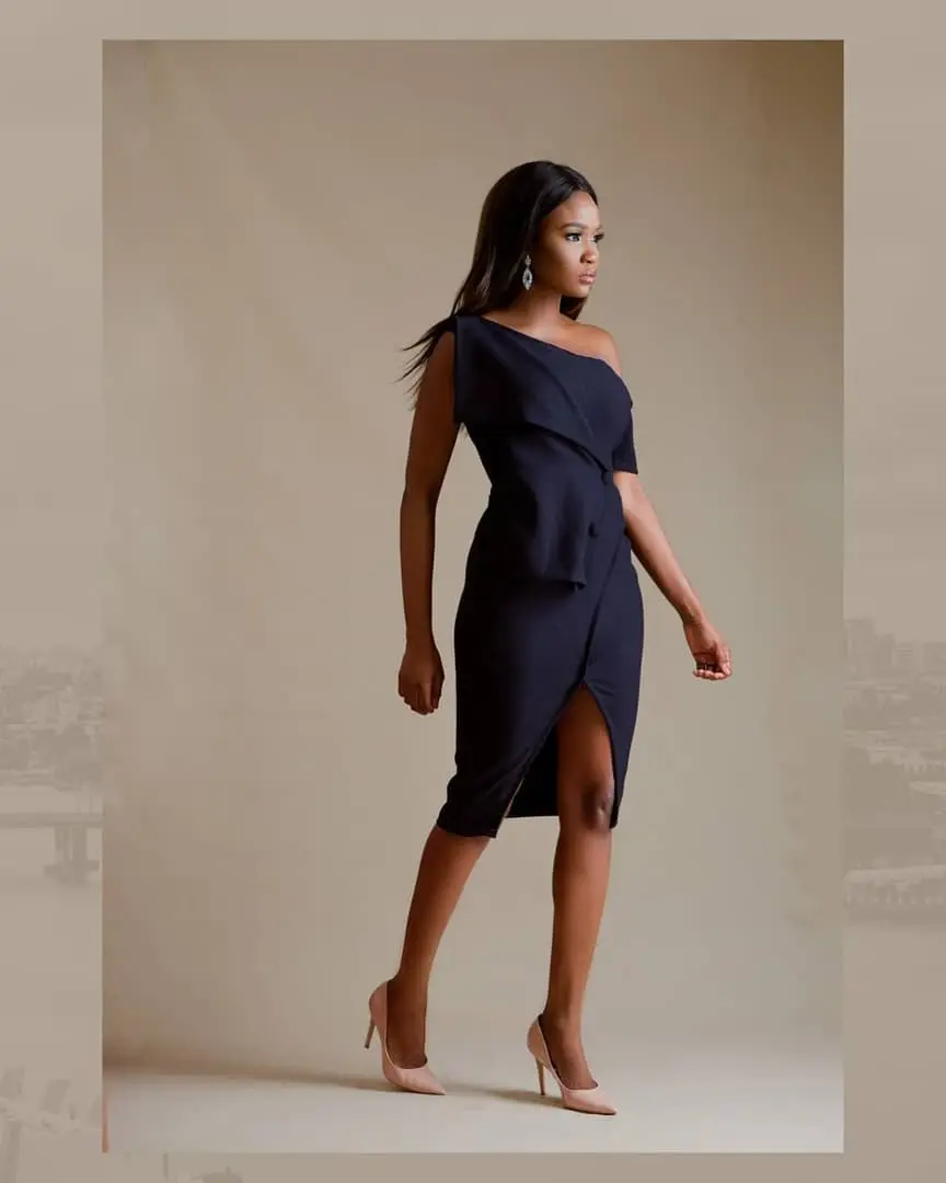 Model mid-stride in an asymmetric navy dress with front slit and blush heels against a beige backdrop.