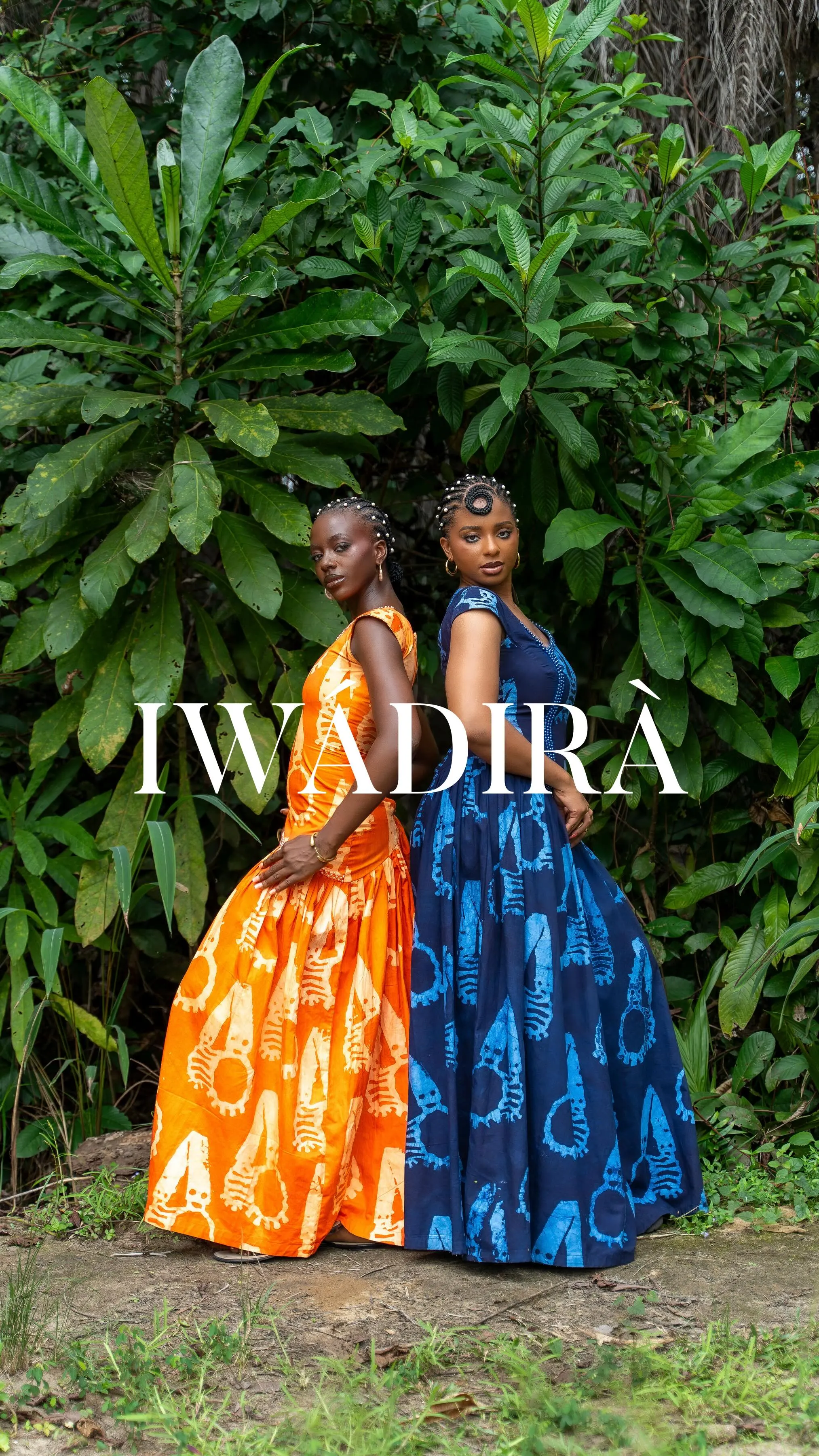 Two models back to back in adire gowns, one orange and one indigo, standing in lush green foliage beneath the Iwadirà wordmark.