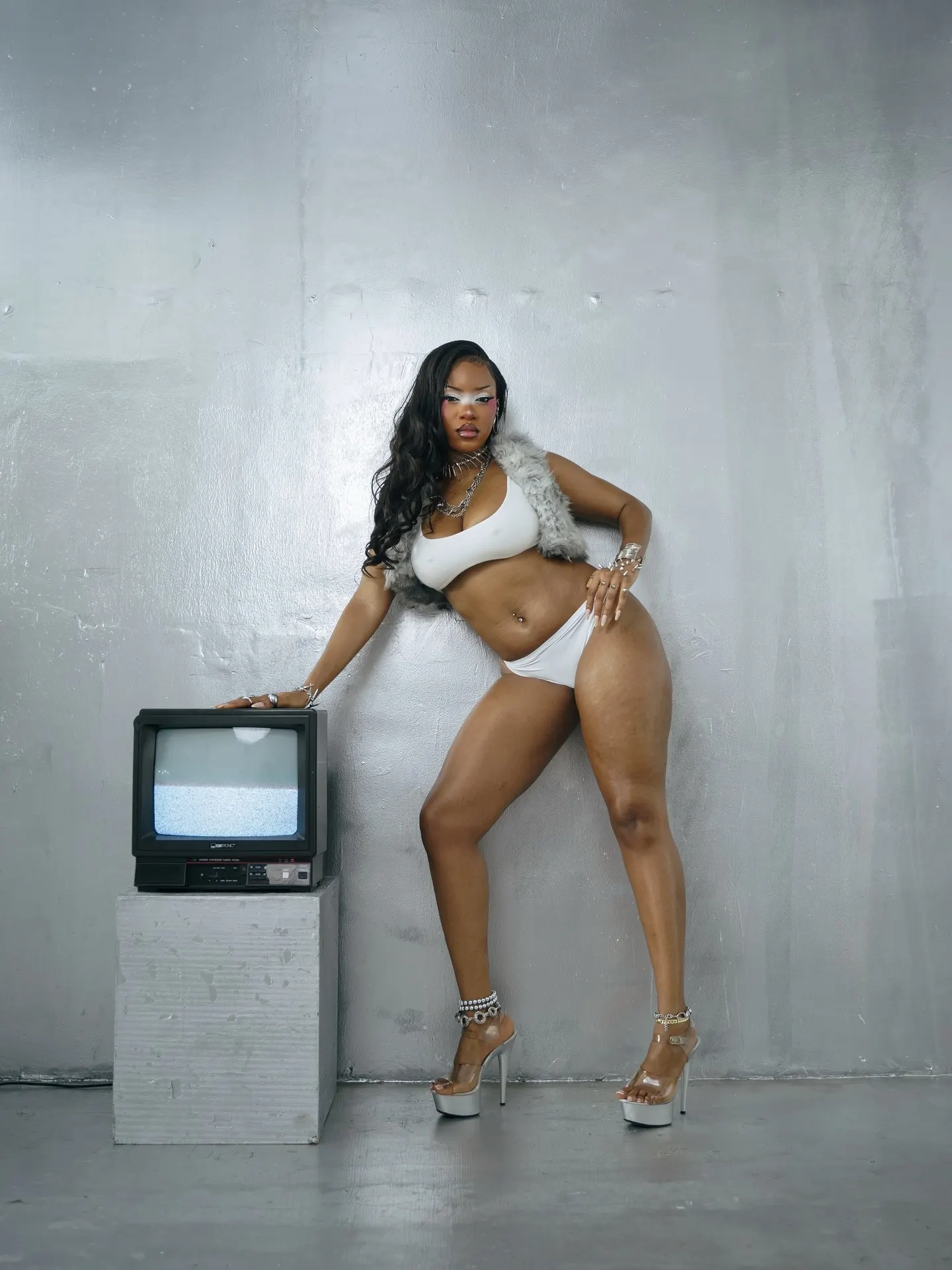 Model in a white bralette and briefs with a grey fur stole and chrome jewelry, standing with one hand on a static-screen CRT television against a silver-foil wall.