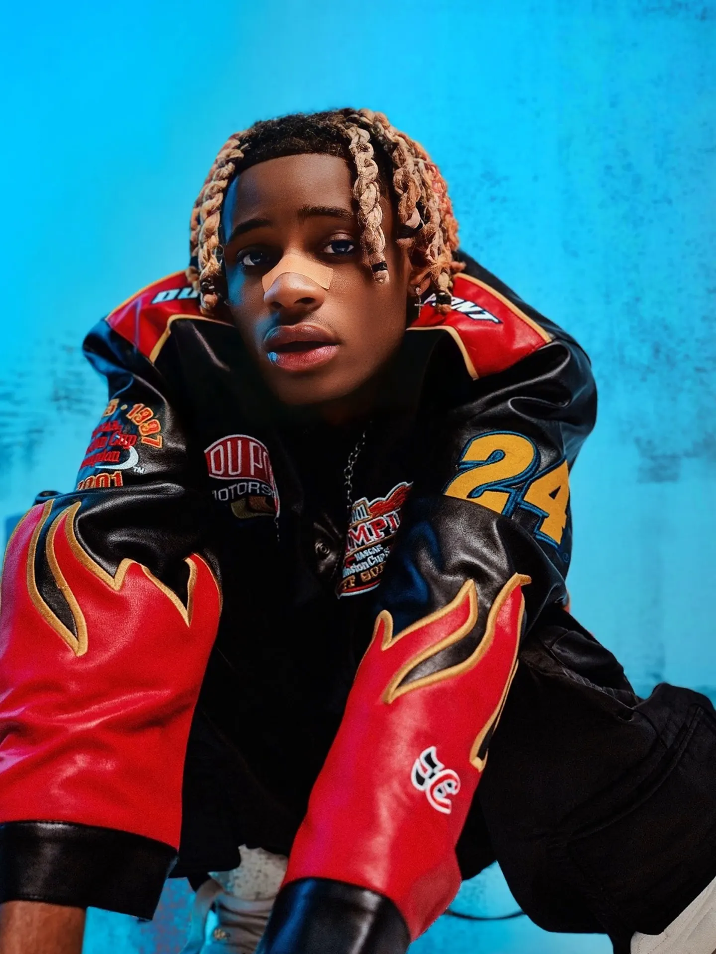 Artist with blond-tipped braids crouches toward the camera in a black racing jacket with red flame-gauntlet sleeves and a yellow 24 patch, against a bright blue backdrop.