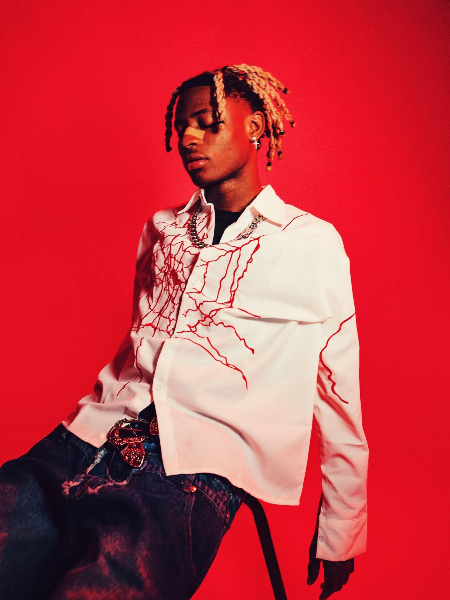 Artist with blond braids leans back with eyes closed in a white shirt embroidered with red thread lines, seated against a saturated red backdrop.