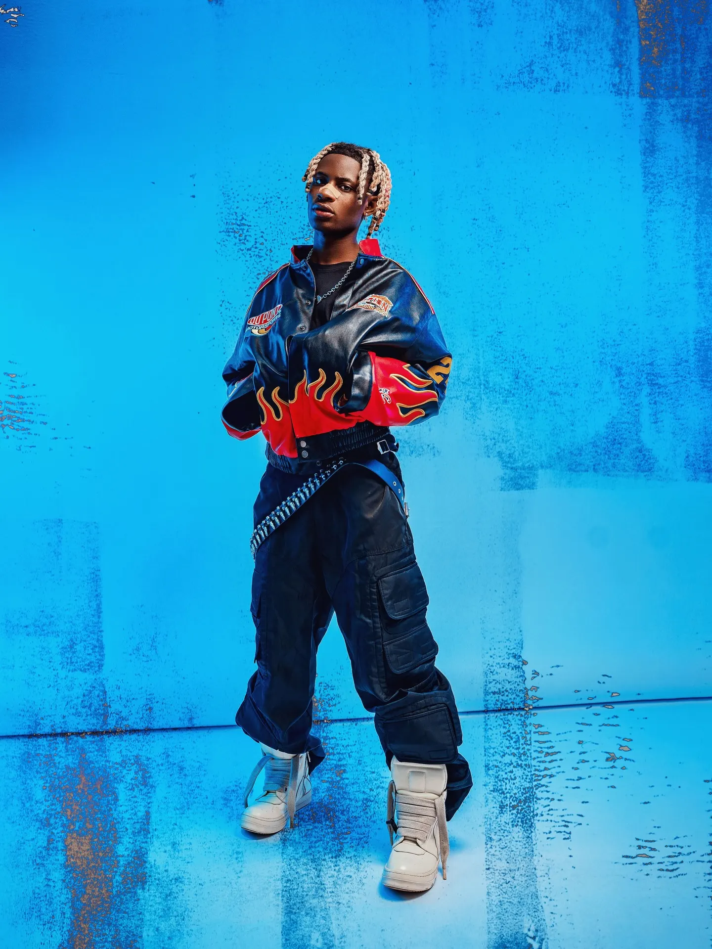 Artist stands full-length with arms crossed in a flame-sleeved racing jacket, black cargo trousers and cream high-top sneakers on a textured blue backdrop.