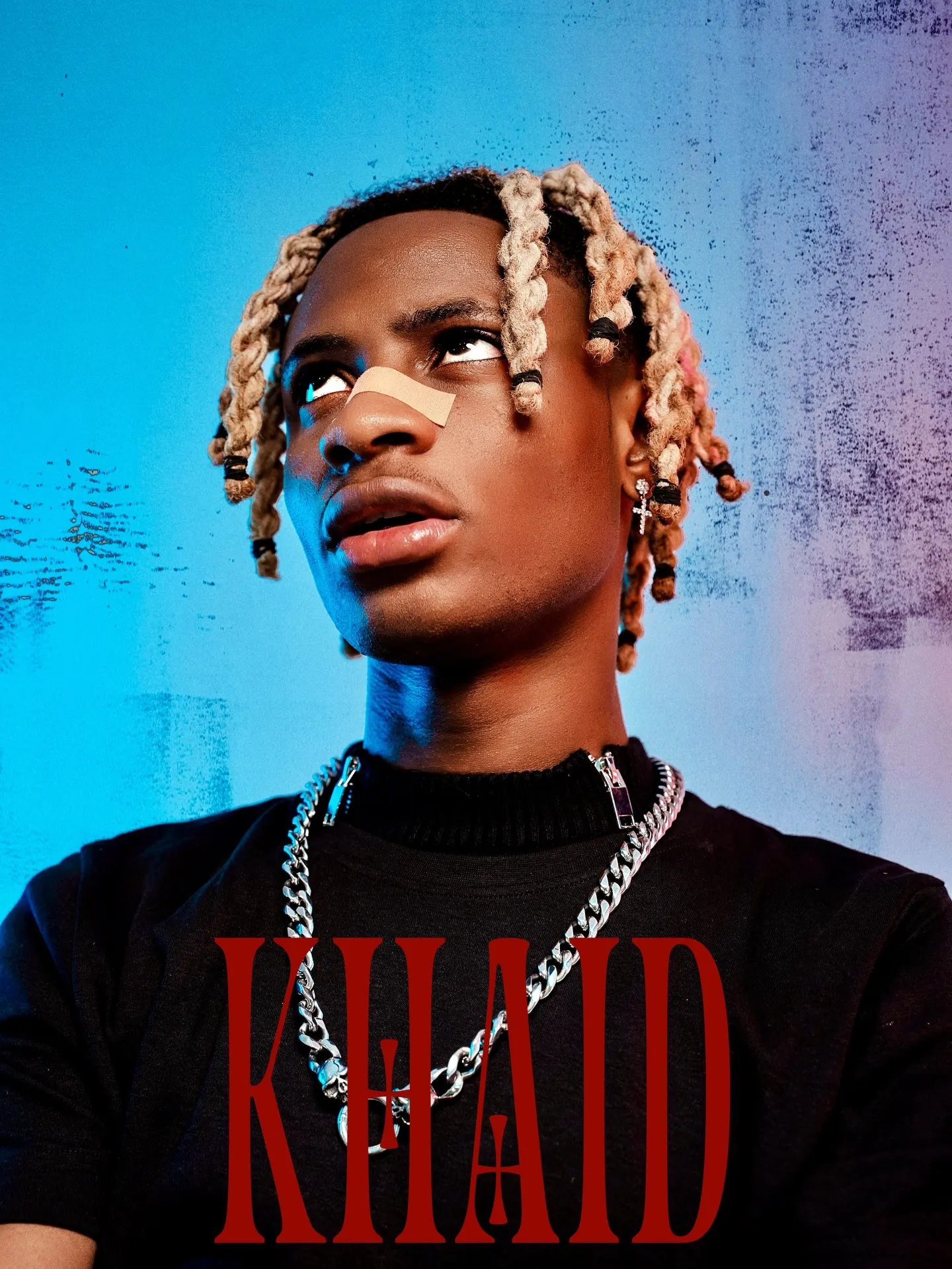 KHAID title card — the artist in a black tee with layered silver chains and blond braids, a plaster across his nose, gazing upward against a scratched blue wall with red type over his chest.