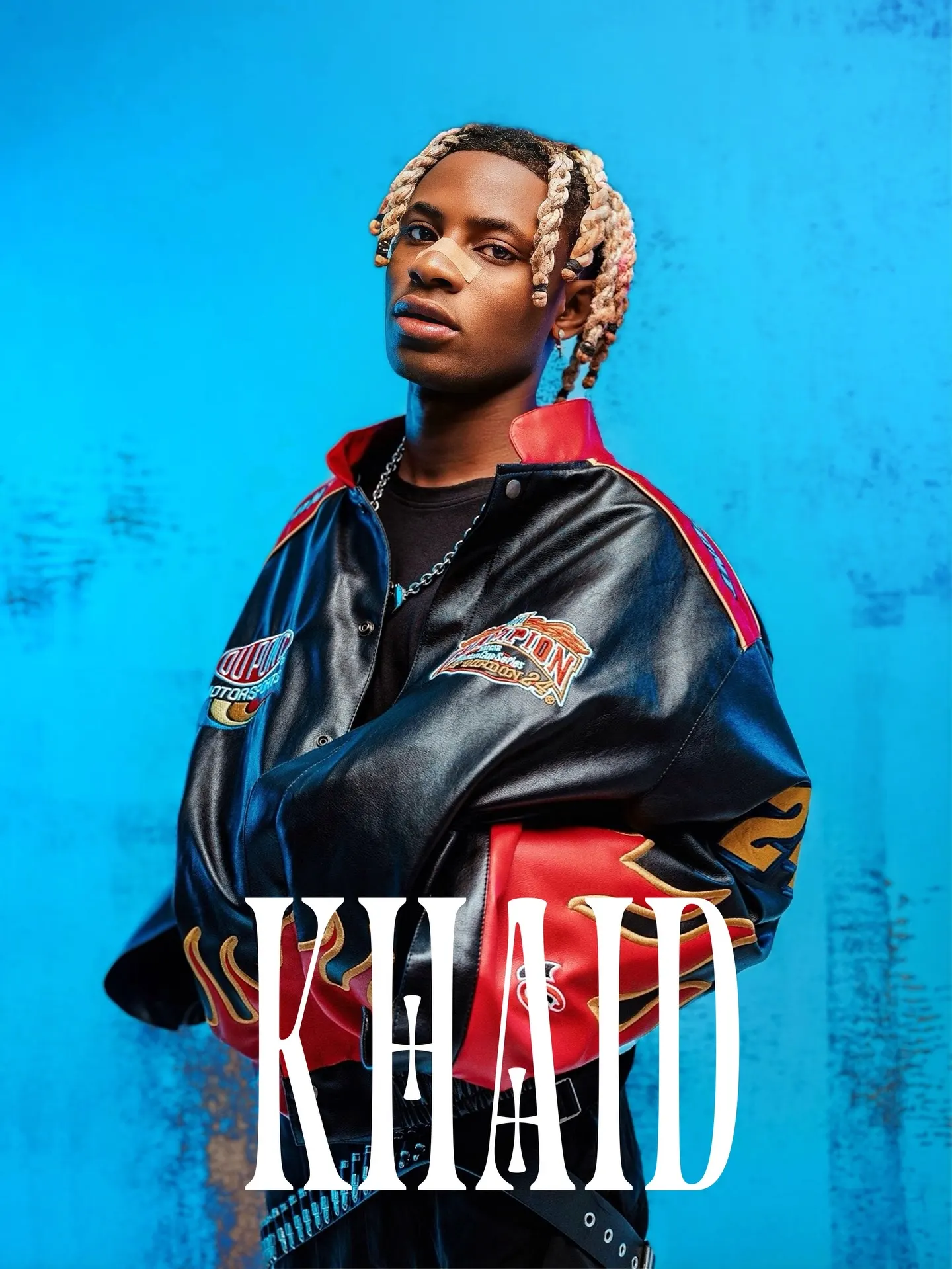KHAID title card — Khaid squared to the camera in an oversized black-and-red racing jacket with flame sleeves, blond braids tied back, white type stacked across a saturated blue backdrop.