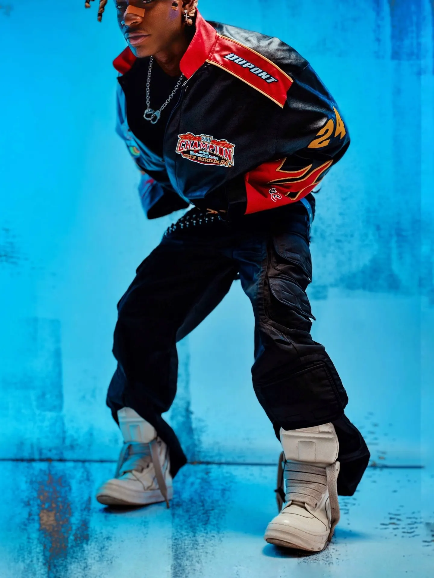 Khaid crouched low in the flame-sleeved racing jacket, black cargo trousers and chunky cream high-tops, knees bent wide against the mottled blue wall.