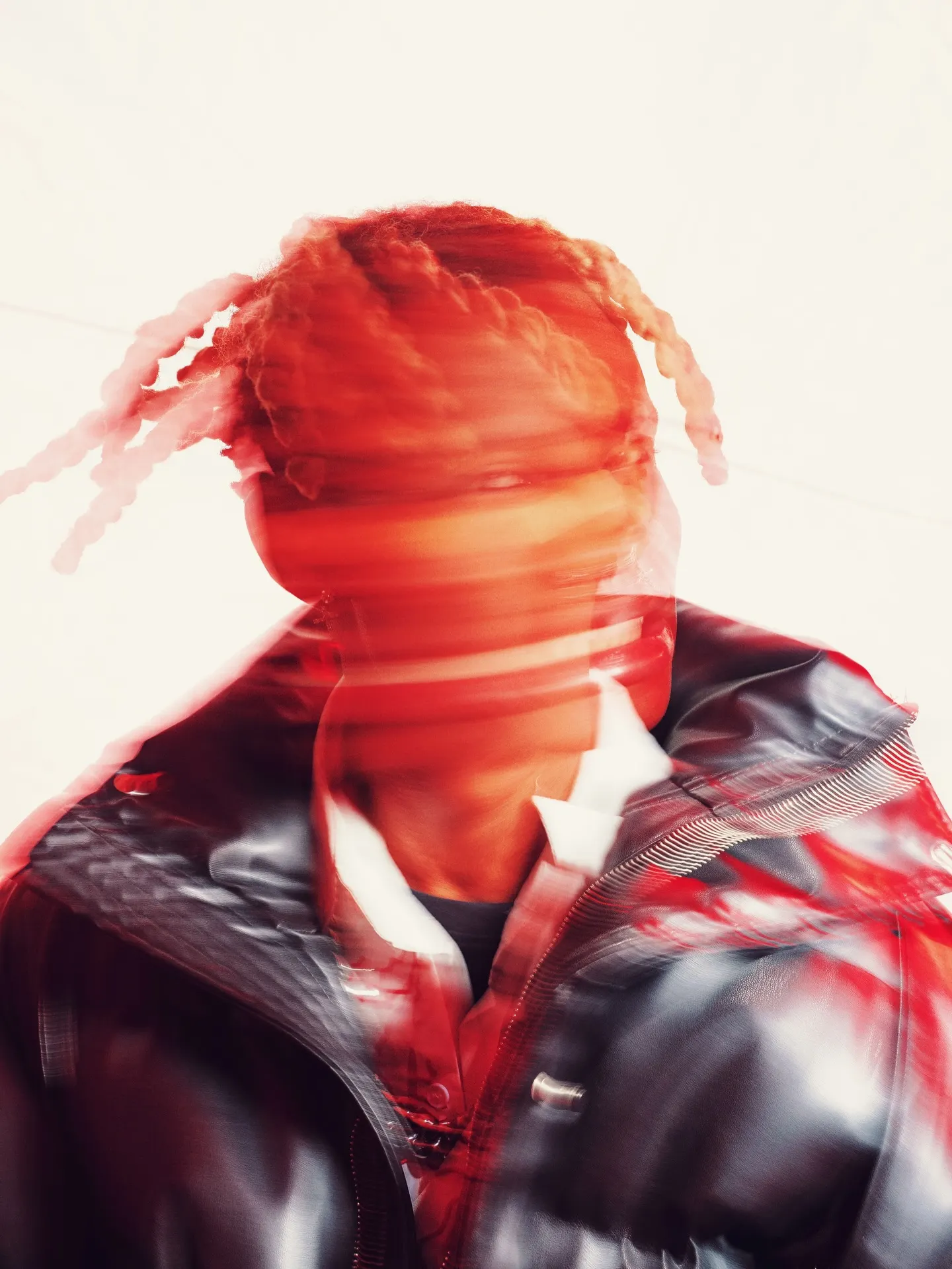 Khaid's braids and face smeared into a streak of red motion blur above a glossy black leather collar, white studio wall behind.