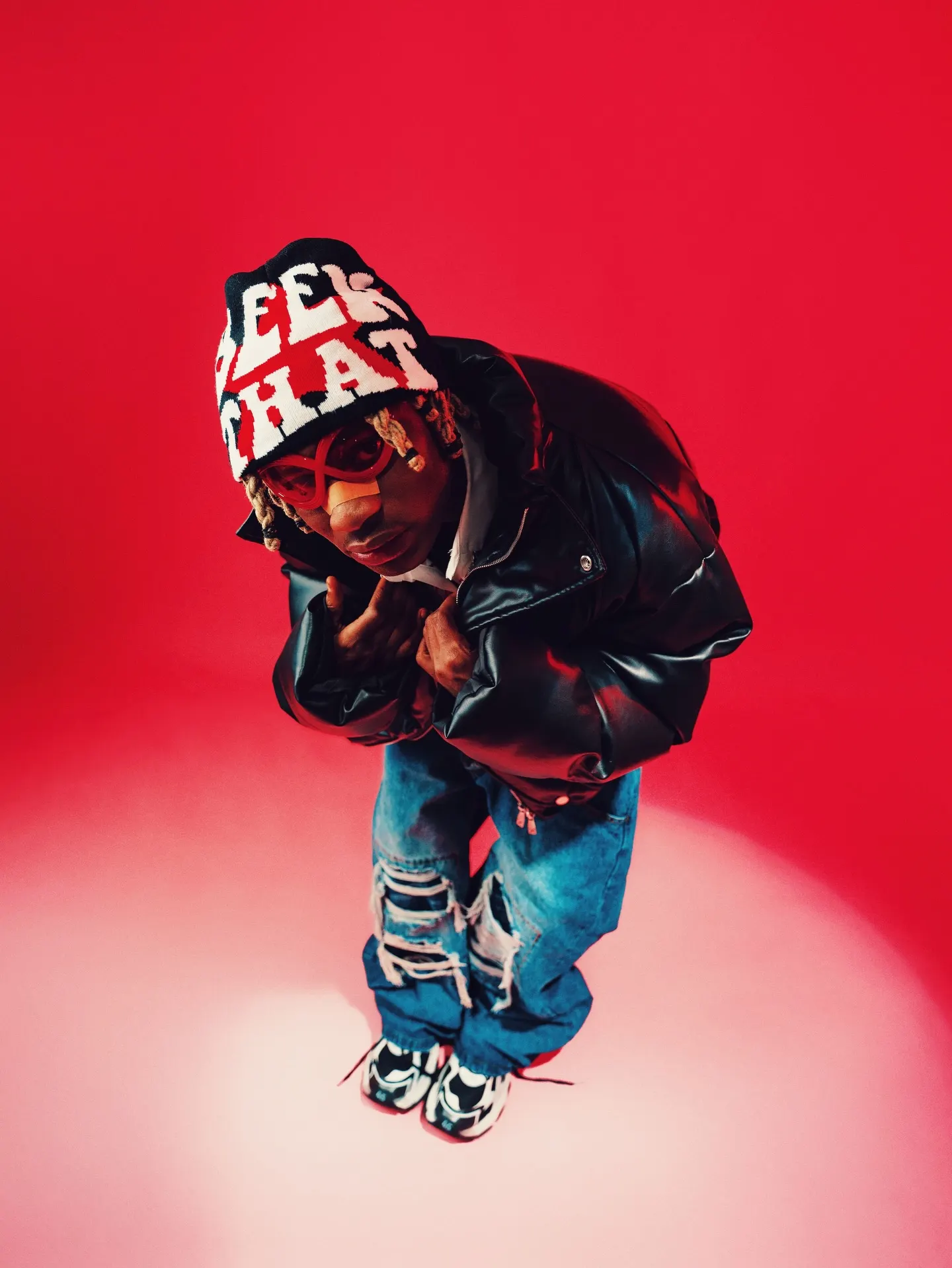 Khaid shot from above in a 'SEEK THAT' beanie and red-tinted shades, hunched forward in a puffy black leather jacket and ripped jeans on a red floor pooled with light.