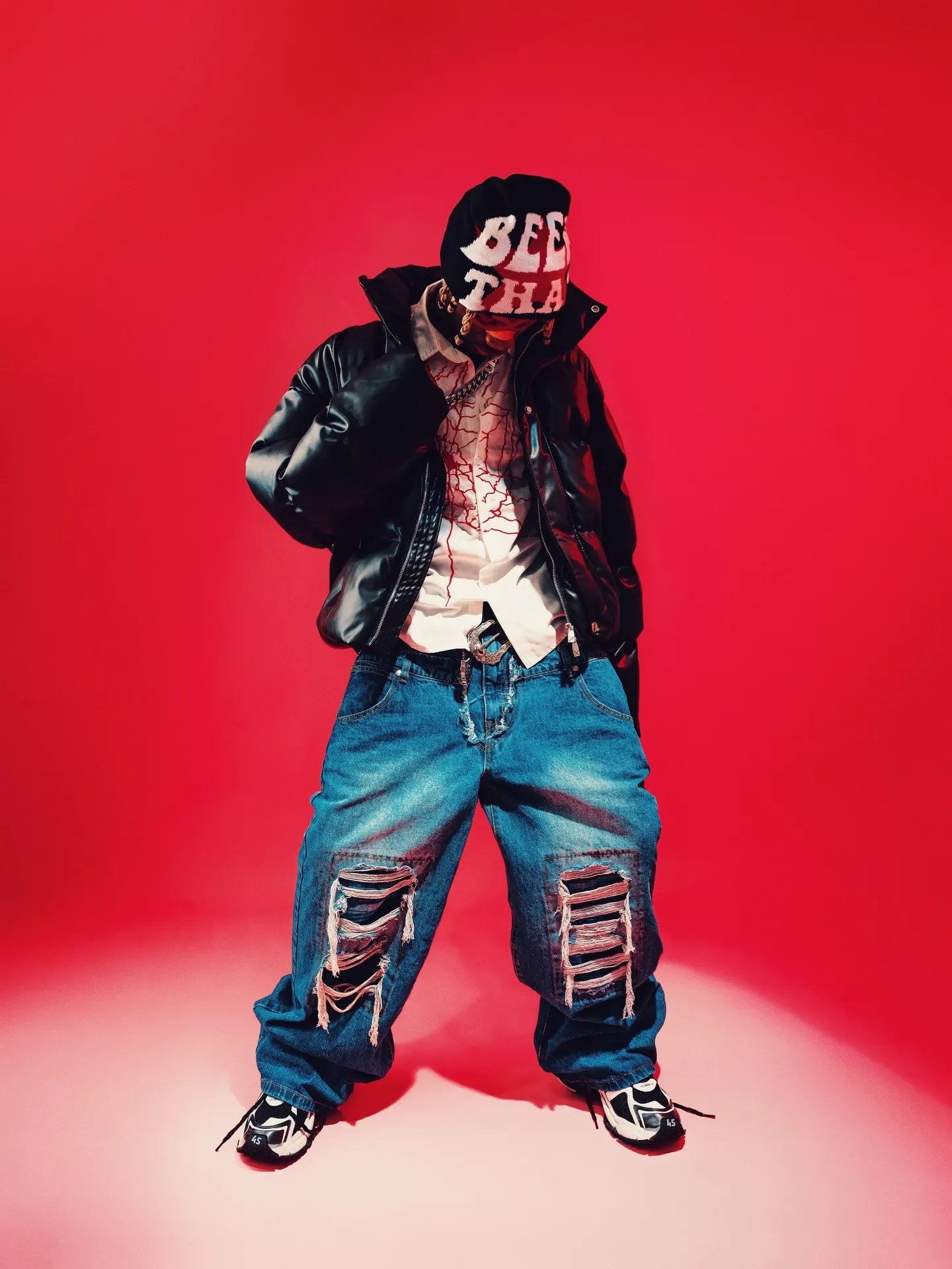 Khaid full-length on red, beanie pulled low over his face, black leather jacket open over the embroidered white shirt, baggy shredded jeans stacked over black-and-white sneakers.