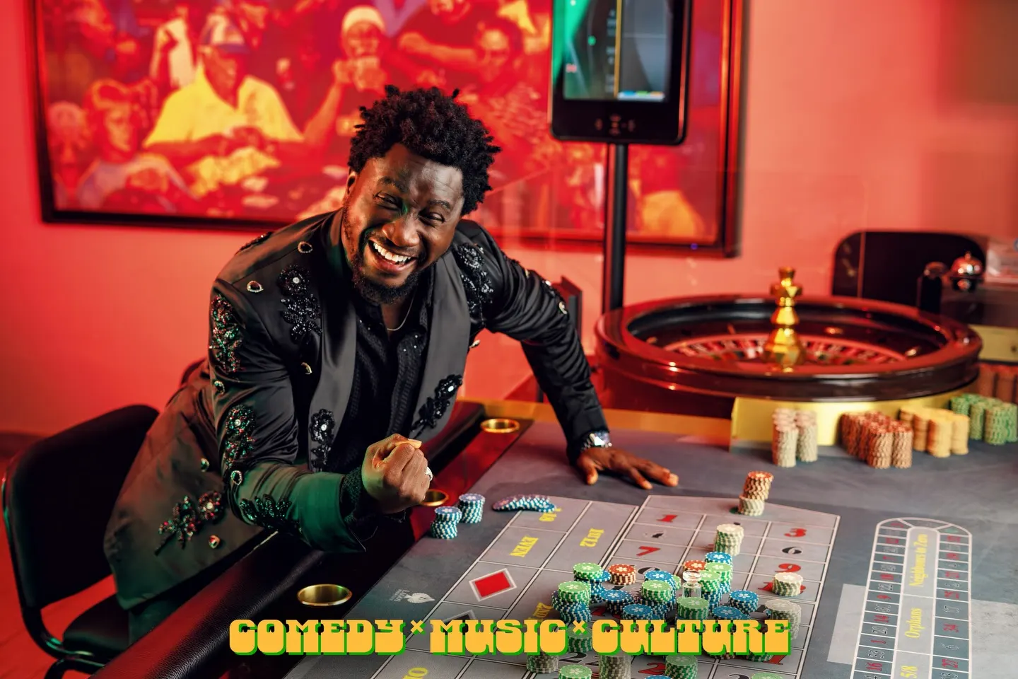Man in a black jacket embroidered with beaded florals laughs as he leans across a roulette table stacked with chips in a red-lit gaming room.
