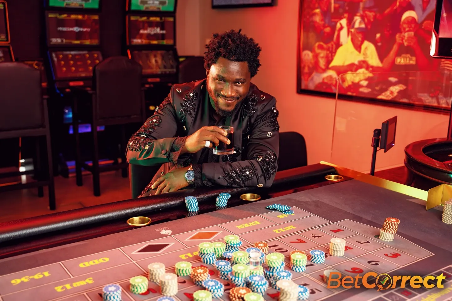 Nasboi sipping a glass over a roulette table crowded with chip stacks, embroidered black suit catching the casino's red arcade light.