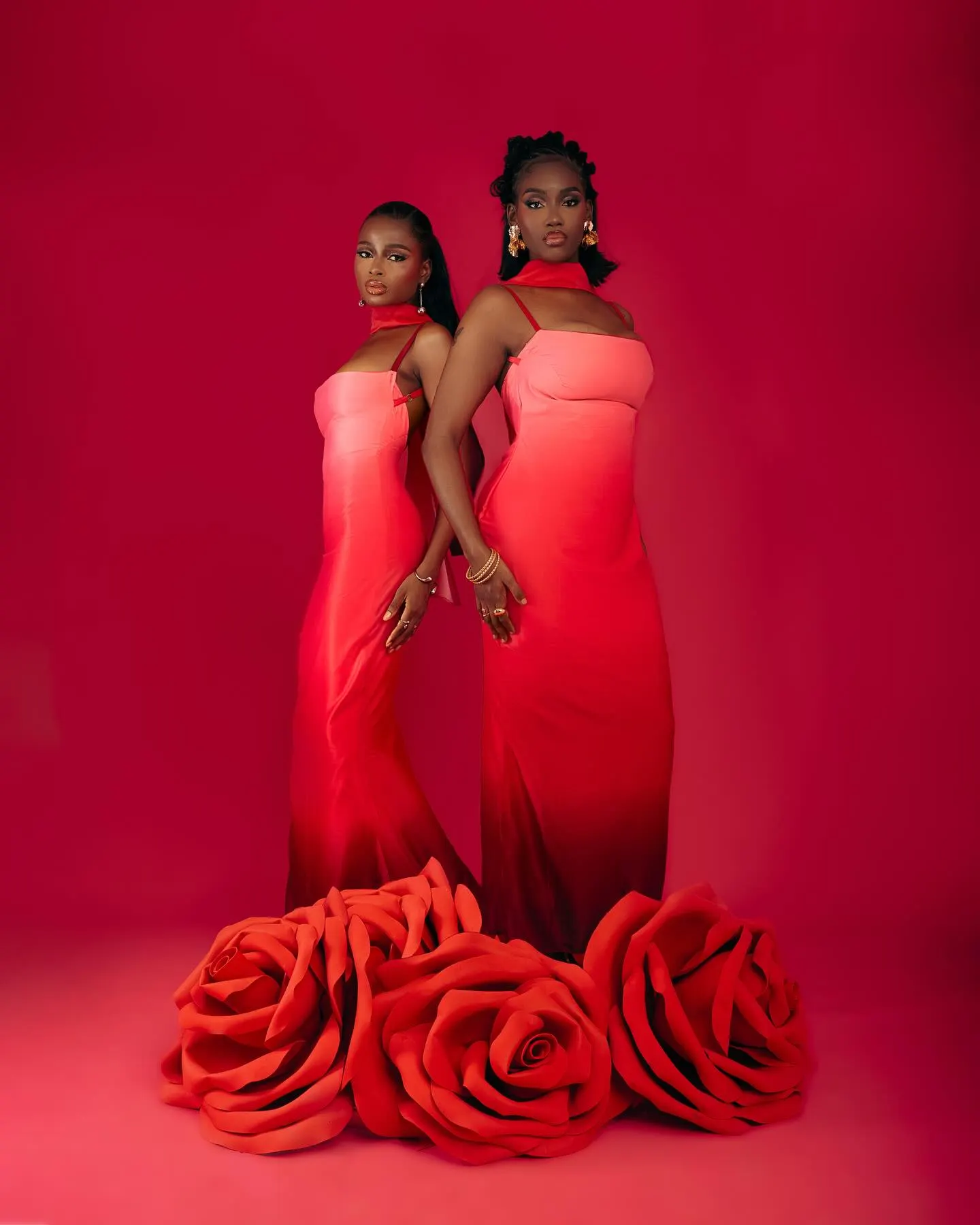 Two models back to back in pink-to-crimson ombré column gowns, rising from a bed of giant red roses against a red backdrop.