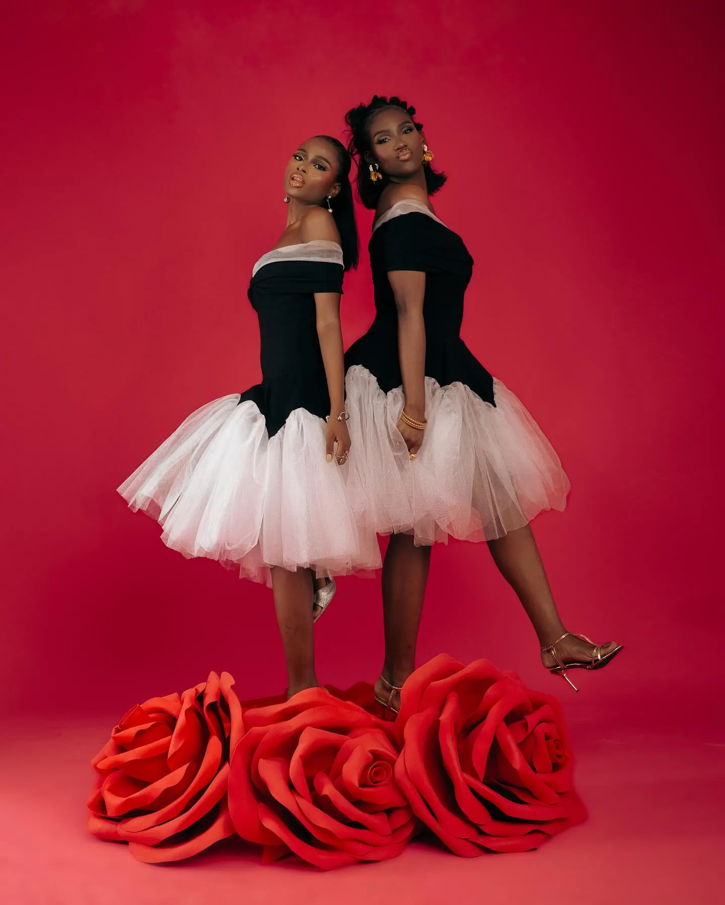 Two models in off-shoulder black bodices over white tulle skirts, one heel raised mid-step atop oversized red roses against a red backdrop.