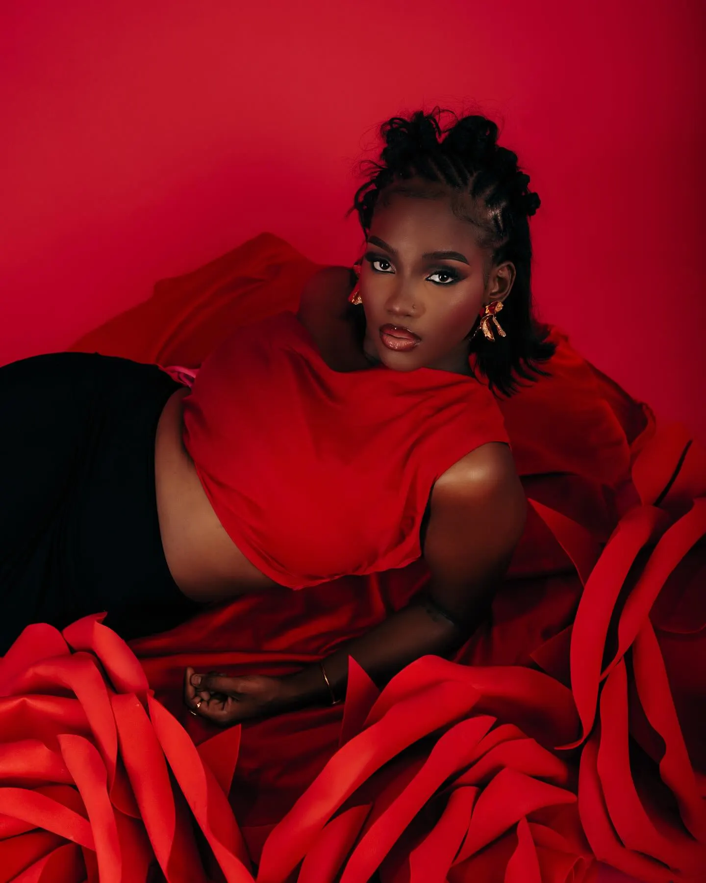 Model in a red draped top reclining across a sea of sculpted red rose petals, gaze fixed on the camera.