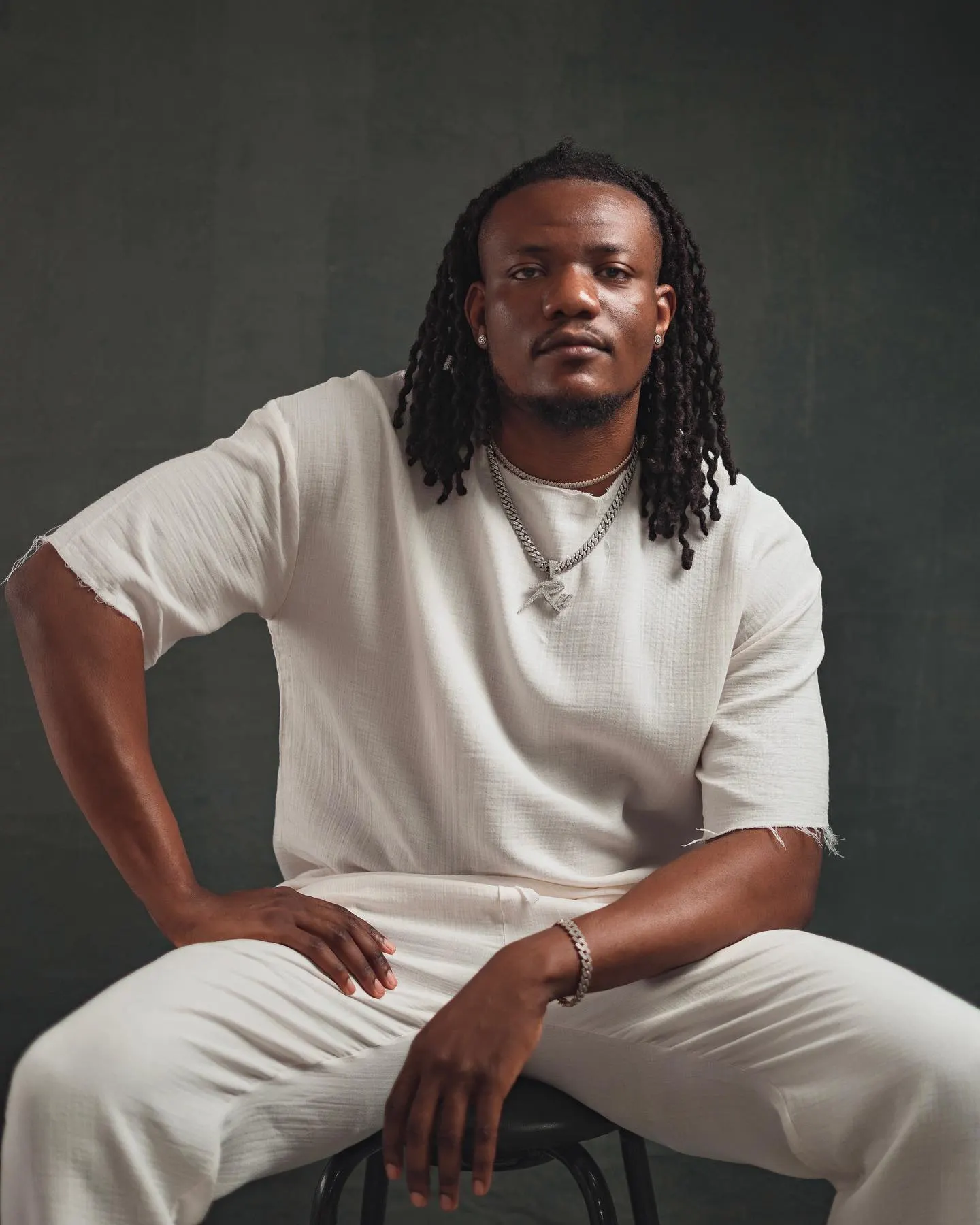 Musician with shoulder-length locs in a raw-hemmed white cotton two-piece and silver chains, seated with elbows on knees against a dark green canvas backdrop.