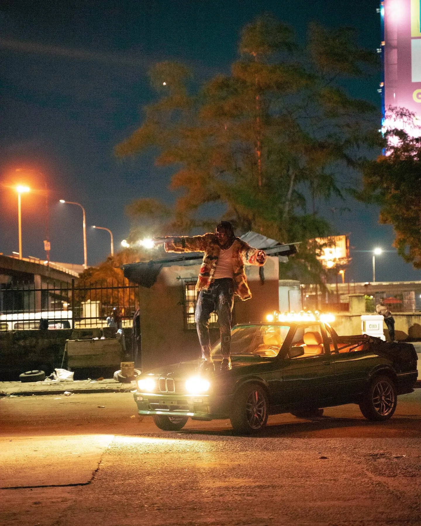 Pheelz standing arms outstretched on the hood of a green convertible at night under streetlights, headlights blazing beneath his feet.
