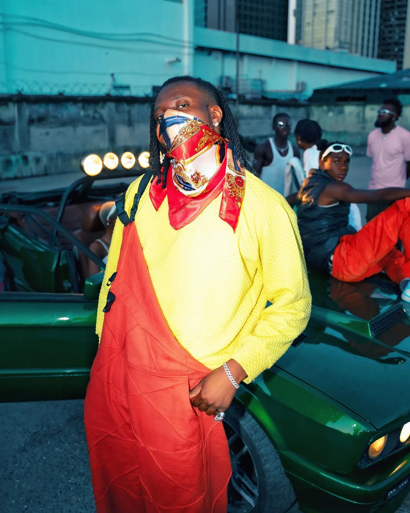 Pheelz in a yellow sweater and orange overalls, face wrapped in a red-and-gold silk scarf, hand in pocket beside a green convertible with friends lounging behind.