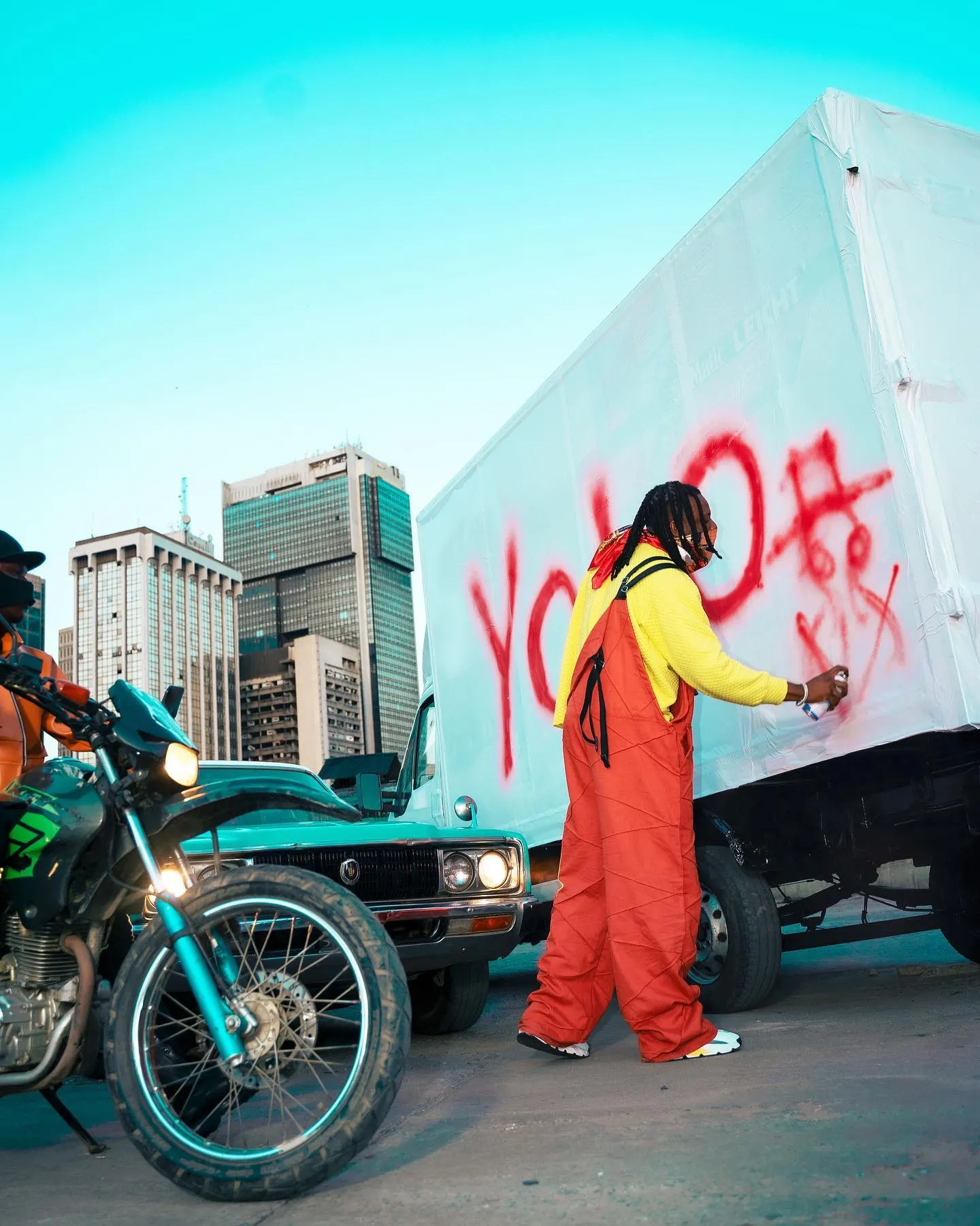 Pheelz in orange overalls spray-painting YOLO in red across a white box truck, motorbike in the foreground and downtown Lagos towers behind.