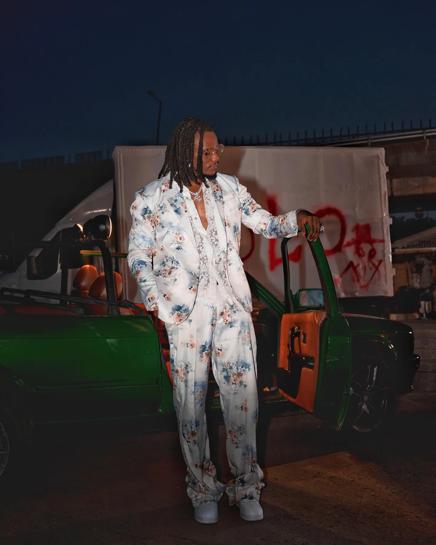 Pheelz in a white floral suit resting an arm on the open door of a green convertible at night, YOLO graffiti scrawled on the white truck behind him.
