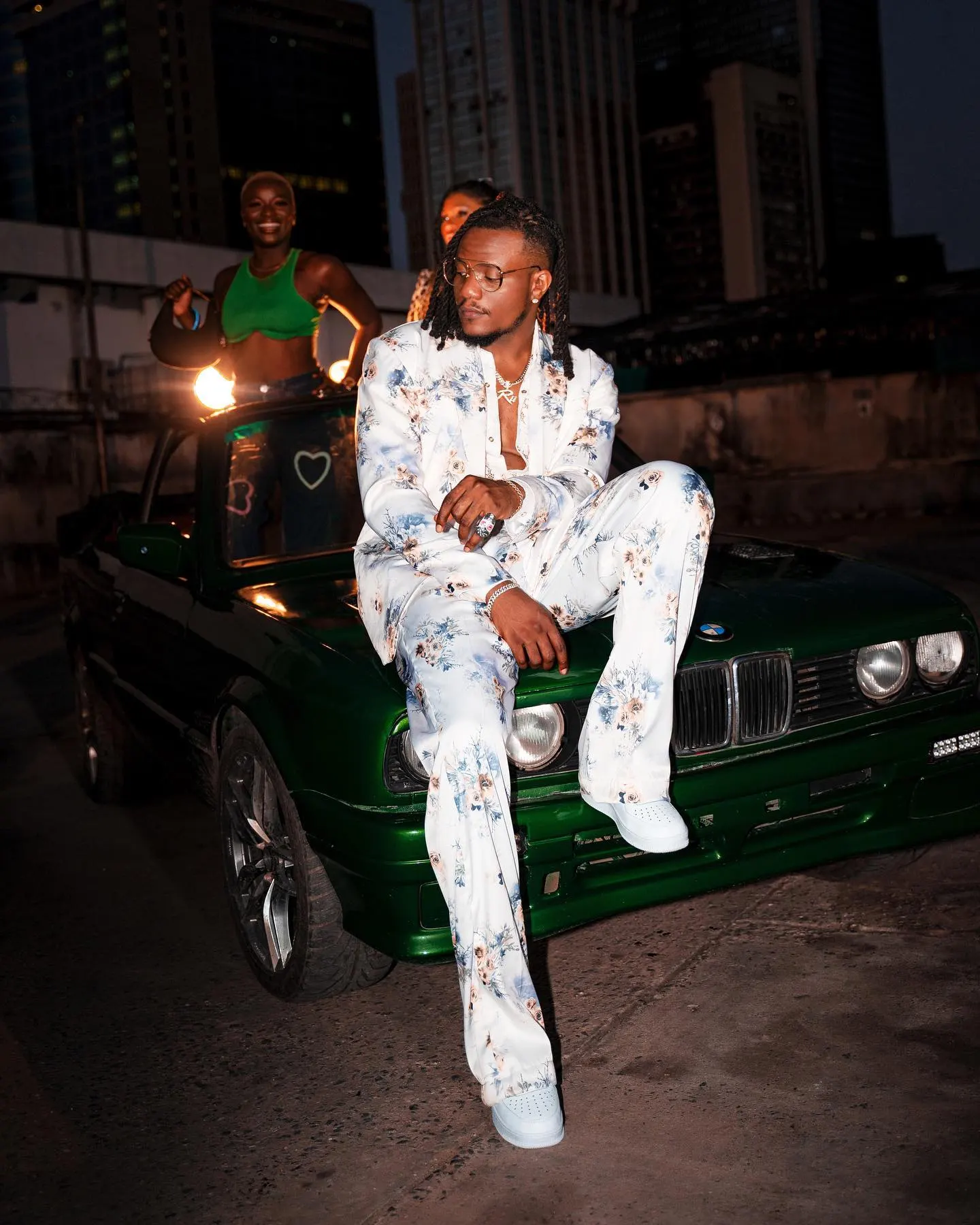 Musician in a white floral suit perched on the hood of a green BMW convertible at night, city towers rising behind him.