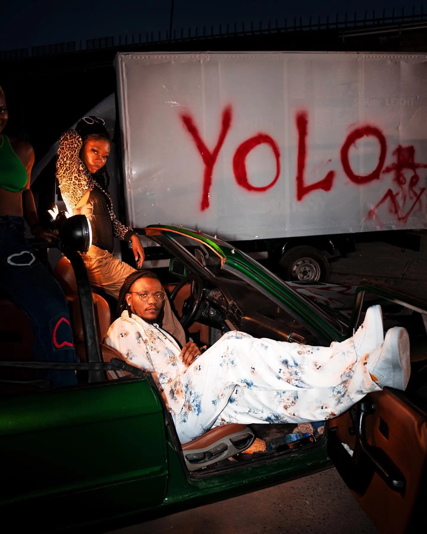 Musician in a yellow knit sweater and orange overalls, face wrapped in a red silk scarf, standing before a green convertible and onlookers in cool daylight.