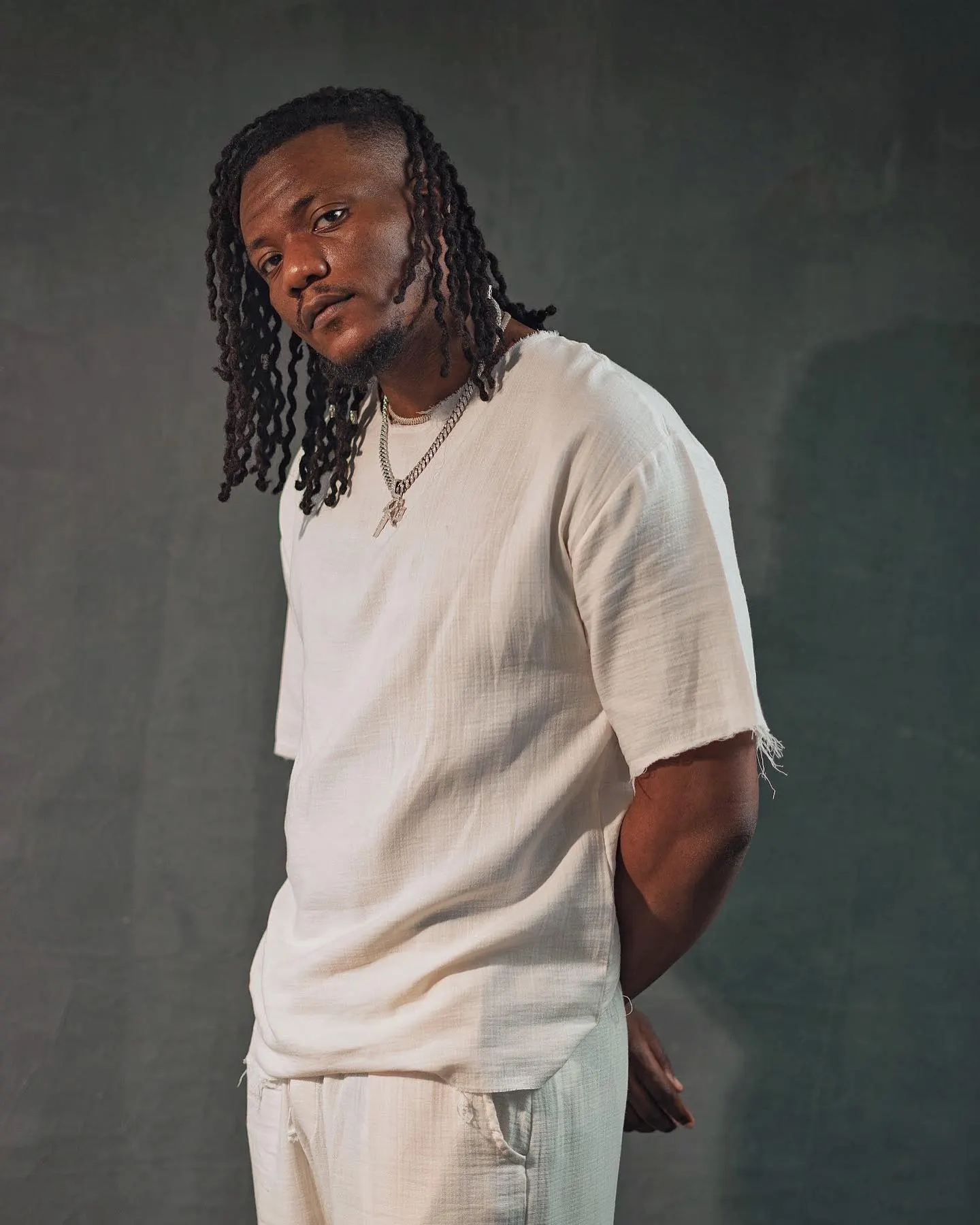 Pheelz in a loose cream two-piece with layered silver chains, hands behind his back, glancing over his shoulder against a mottled grey studio wall.