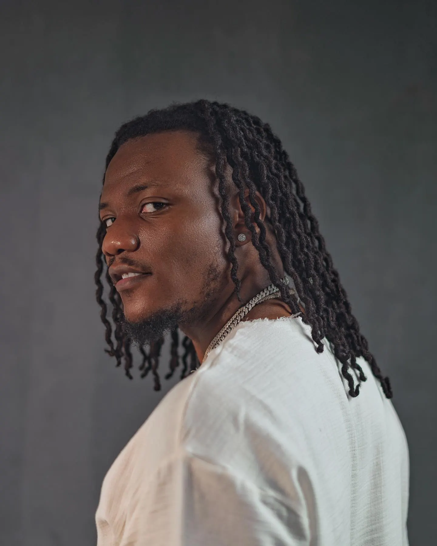 Pheelz in profile, long dark locs falling past his shoulders over a white top, looking back with a soft smile against a grey studio wall.