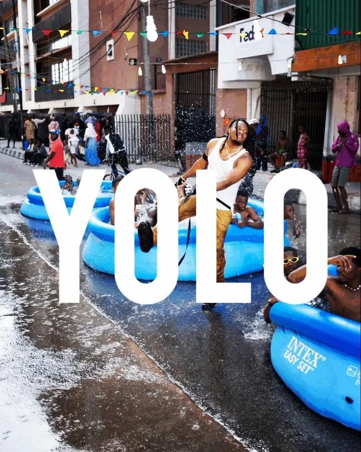 YOLO title card — huge white letters over Pheelz dancing in a white vest between blue inflatable pools full of kids on a wet, bunting-strung Lagos street.