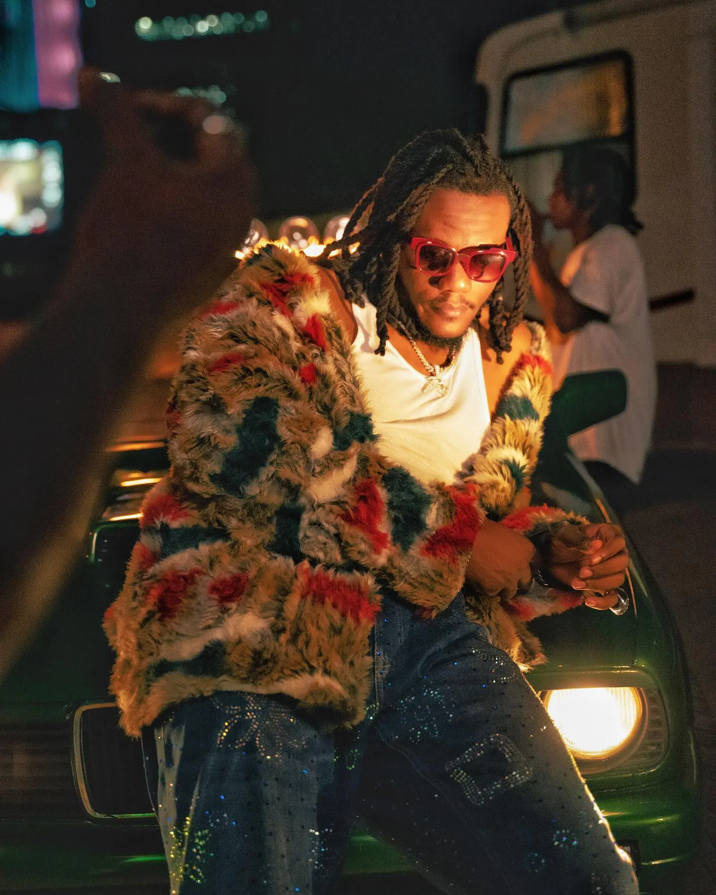 Pheelz in a multicoloured fur coat, white vest and pink shades leaning back on the hood of a green convertible at night, a phone camera framing him from the left.