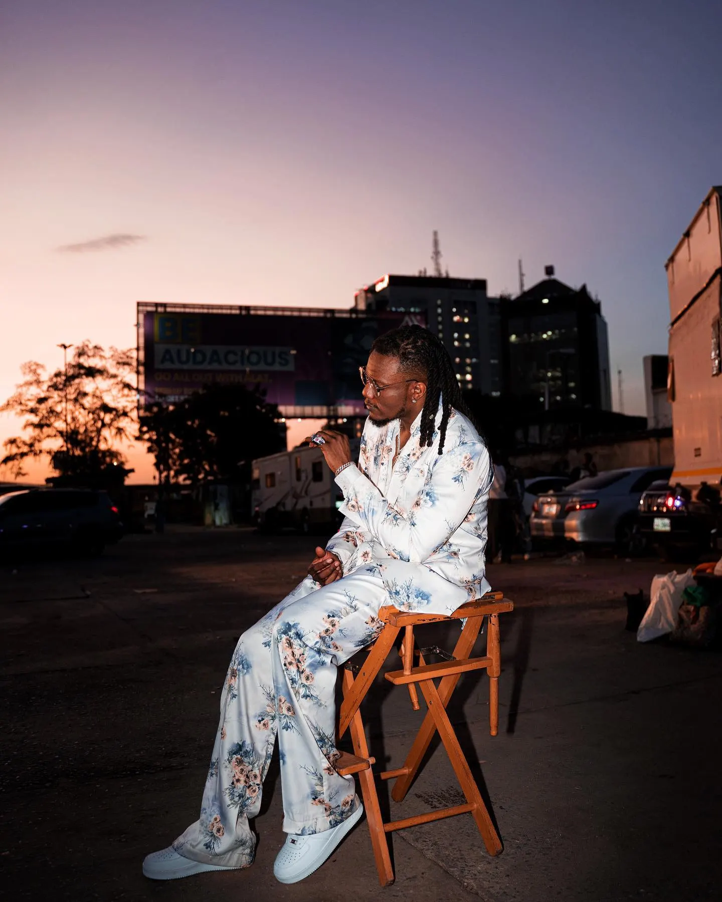 Pheelz in a white floral suit seated on a wooden director's chair in a dusty lot at dusk, city billboard and pink-purple sky behind him.