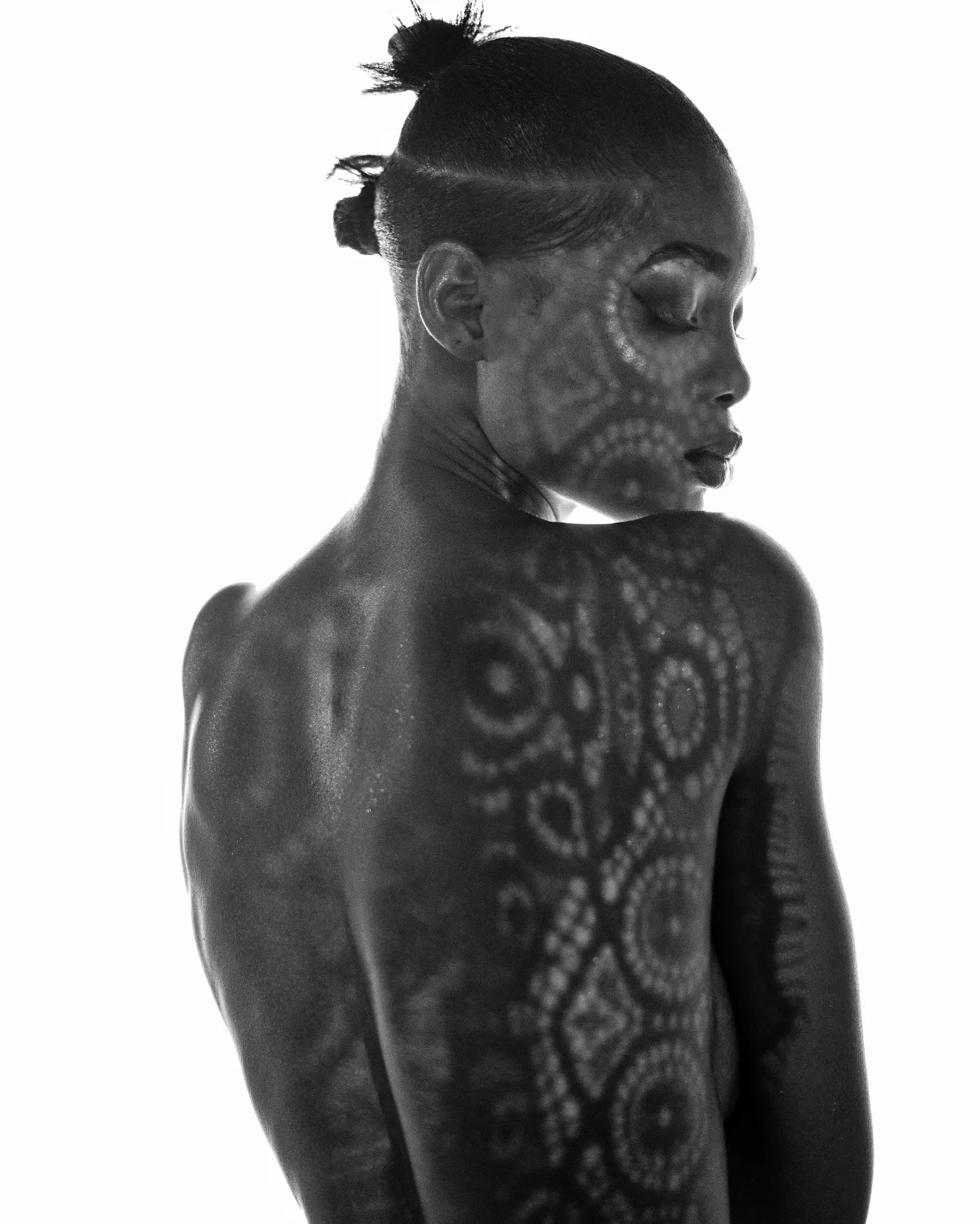 Black-and-white study of a model seen from behind, head turned in profile, circular lace shadows patterning her bare back and face.