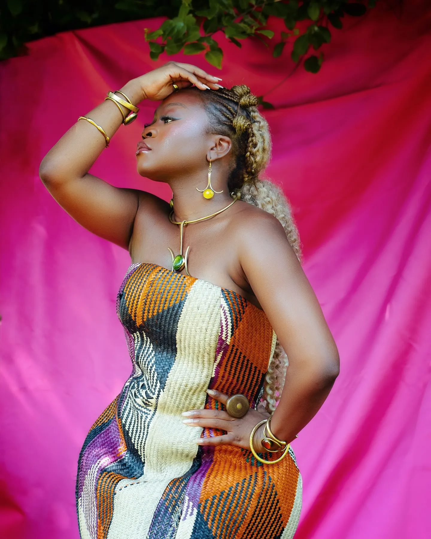 Sampa the Great in a strapless woven raffia dress of orange, cream and plum plaid, one hand raised to her brow, gazing sideways against a hot-pink satin backdrop.
