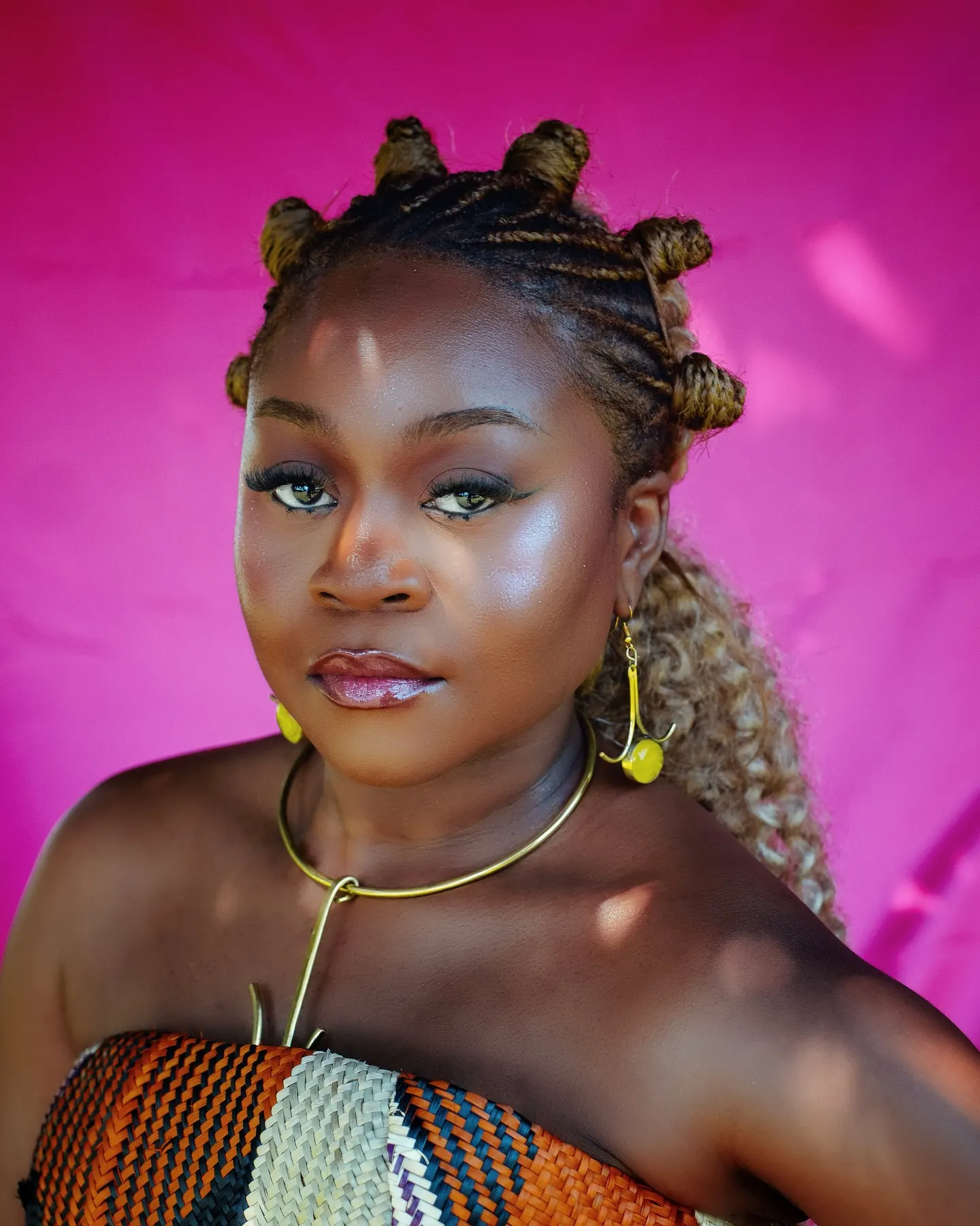 Close portrait of Sampa the Great with bantu knots, gold collar necklace and yellow drop earrings, sunlight dappling her face against a magenta backdrop.