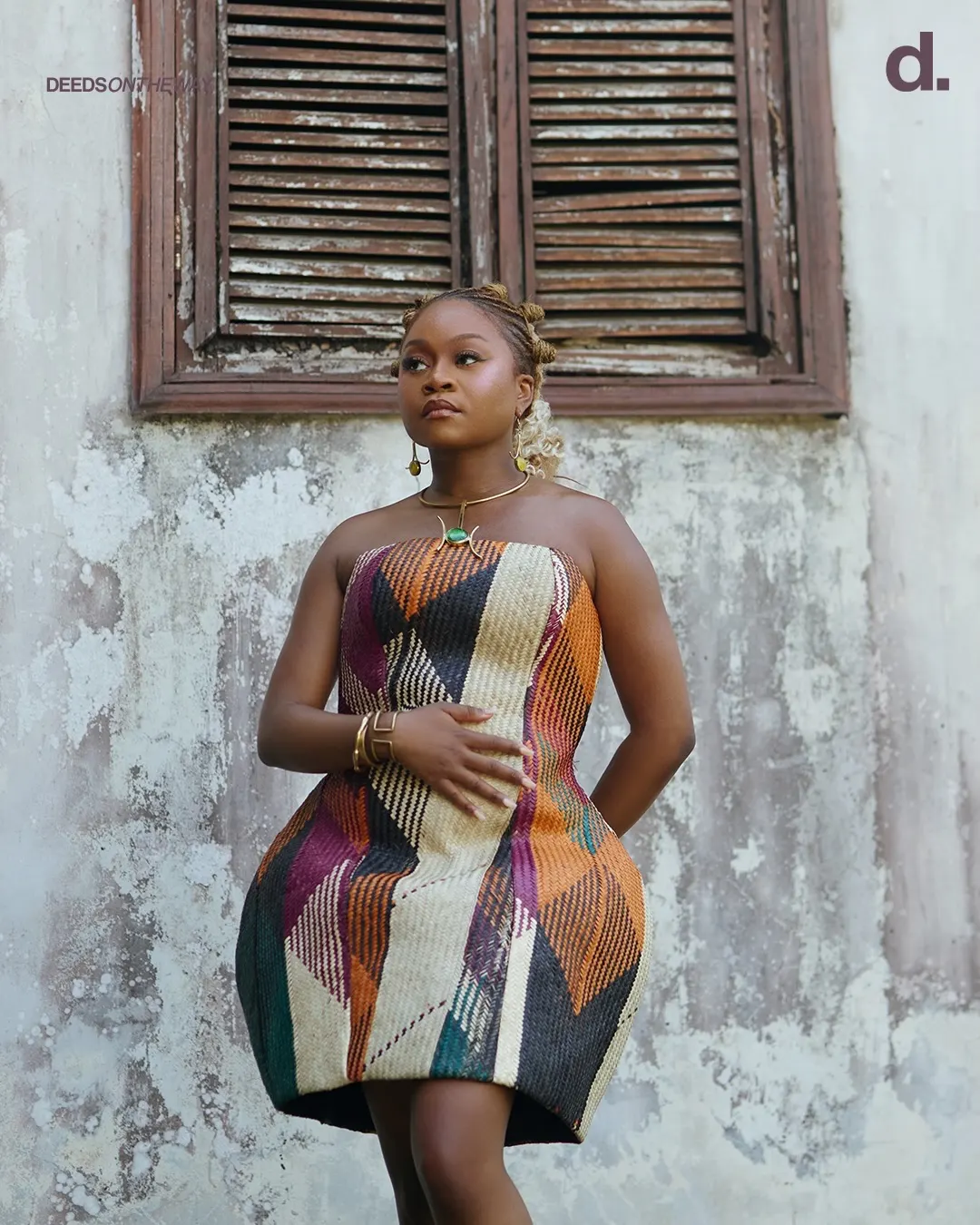 Sampa the Great in a sculptural strapless raffia mini dress, hands at her waist, standing before a weathered plaster wall and dark wooden shutters.