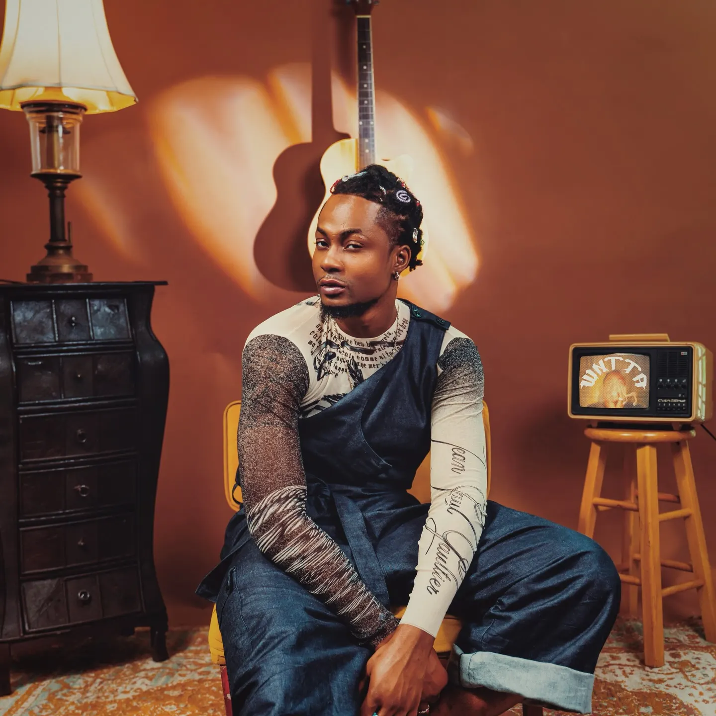 Seyi G in a printed mesh top and denim dungarees leans forward on a yellow chair, an acoustic guitar and vintage TV set against the terracotta wall behind him.