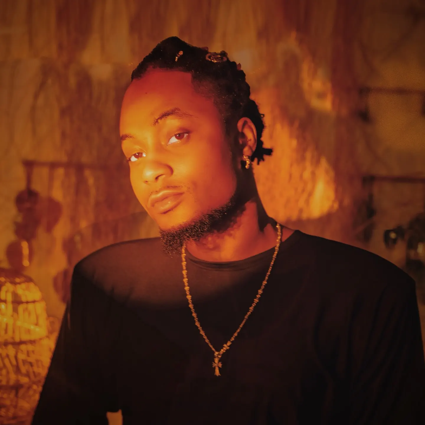 Seyi G in a black tee and long beaded cross pendant, bathed in amber light, glancing over his shoulder against a raffia-textured backdrop.