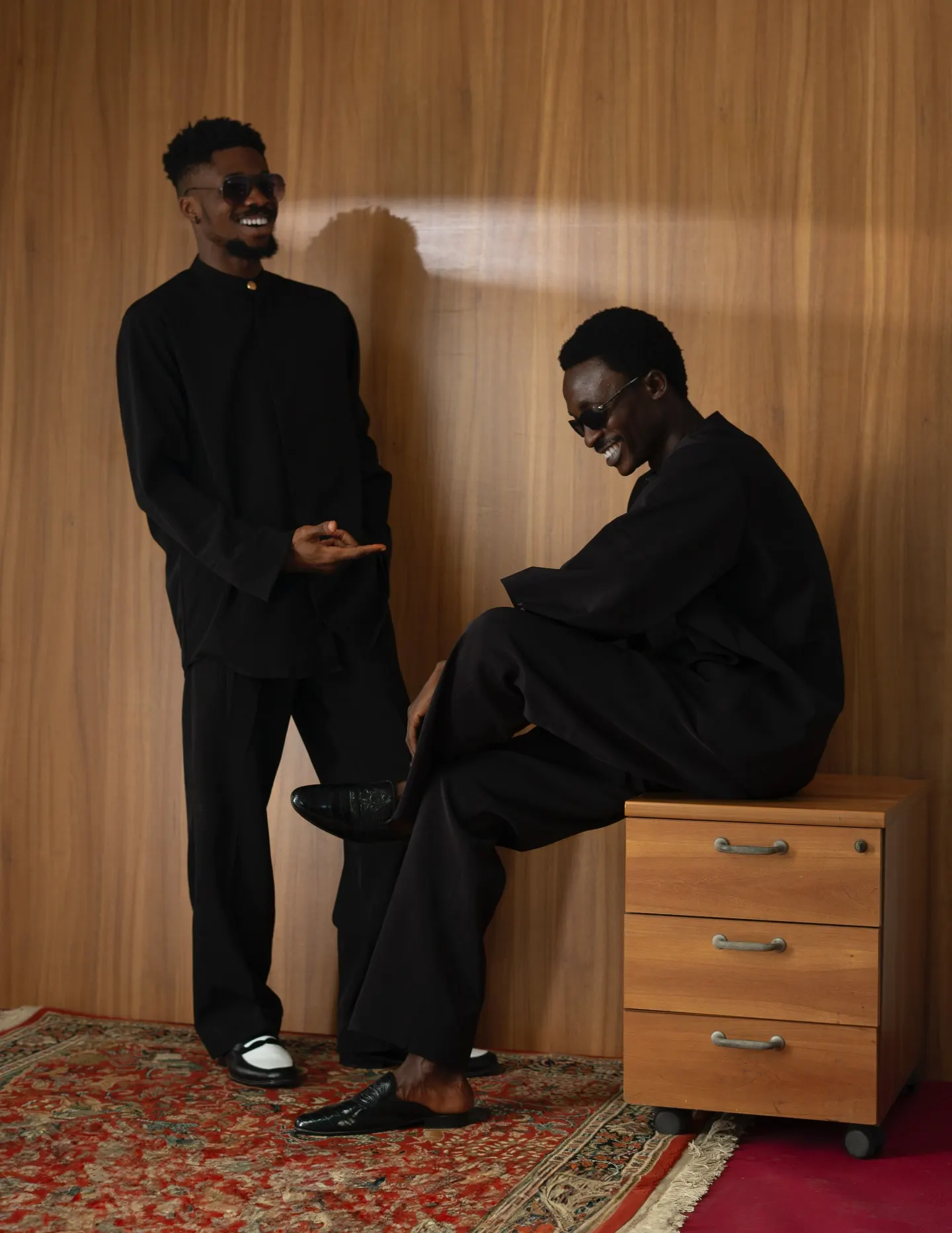 Two models in matching black band-collar suits share a laugh, one perched on a wooden filing cabinet in slippers while the other gestures beside him against a wood-panelled wall.
