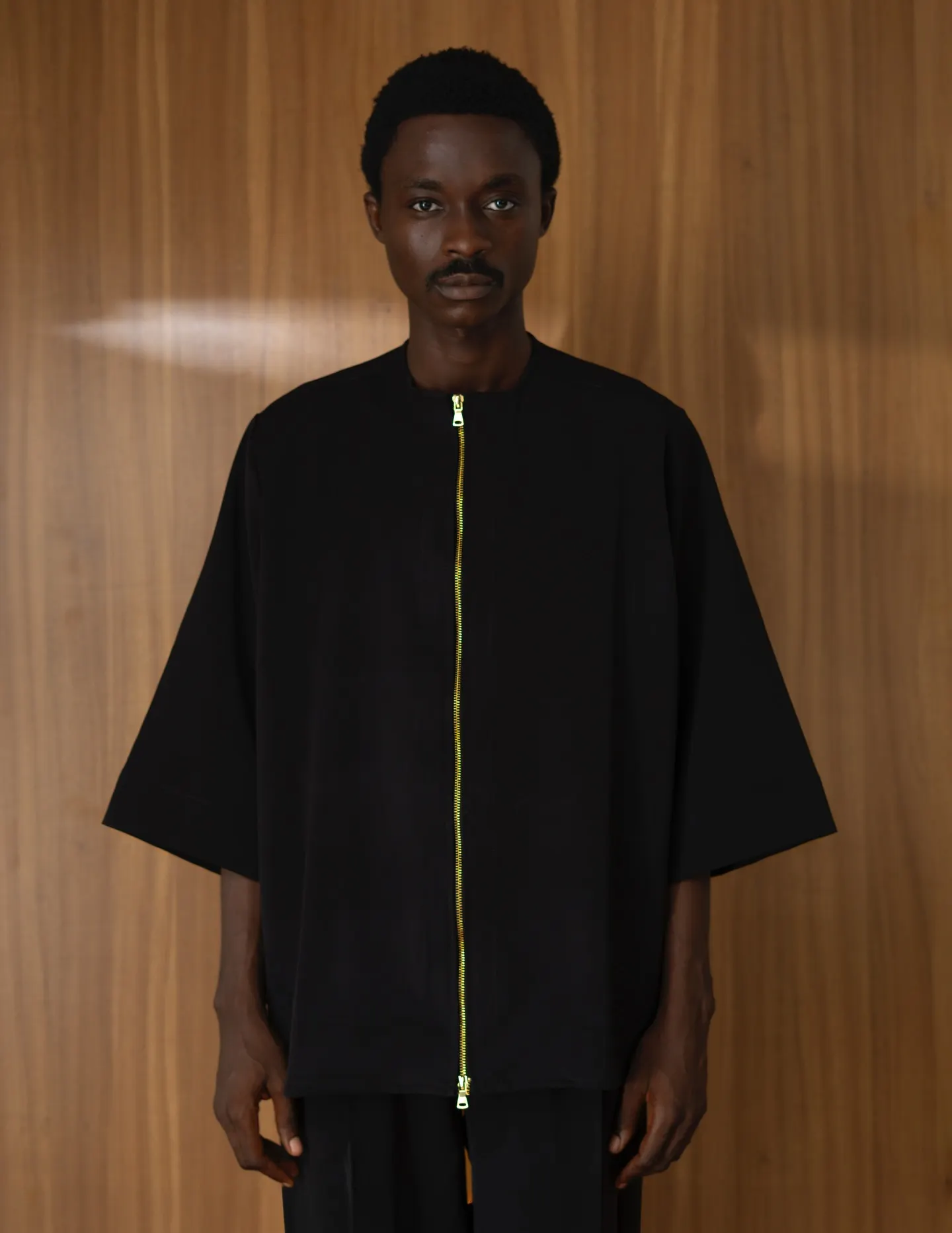 One model tugs the hem of another's green SHOKS tee, seated on a wooden cabinet in a beige tee and dark denim, both atop a Persian rug before a wood-panelled wall.