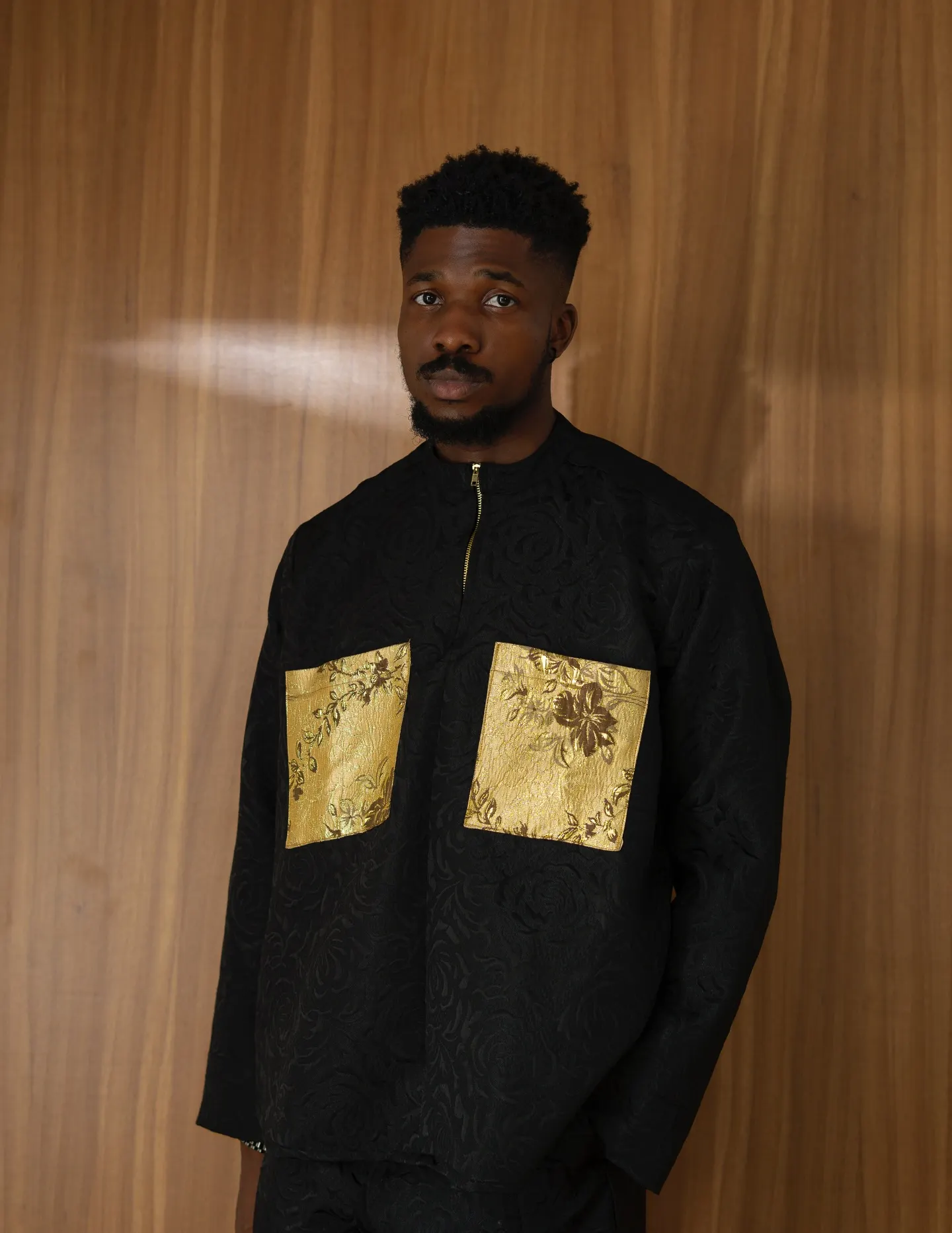 Model in a black jacquard zip tunic with gold brocade patch pockets, gazing at the camera against warm wood panelling.