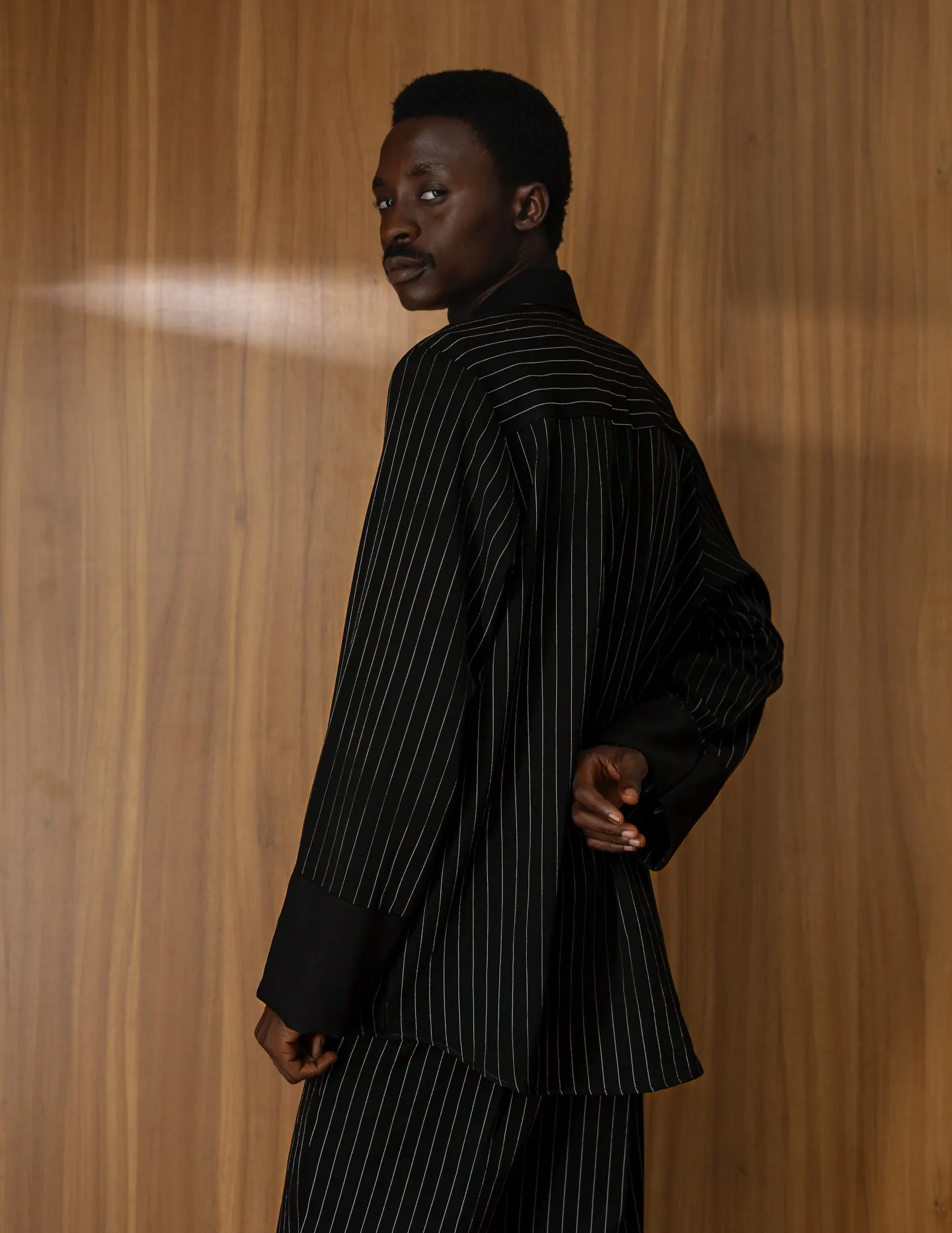 Model in a pinstriped black suit sits atop a small wooden cabinet, one polished loafer propped on his knee, framed by wood panelling and a Persian rug.
