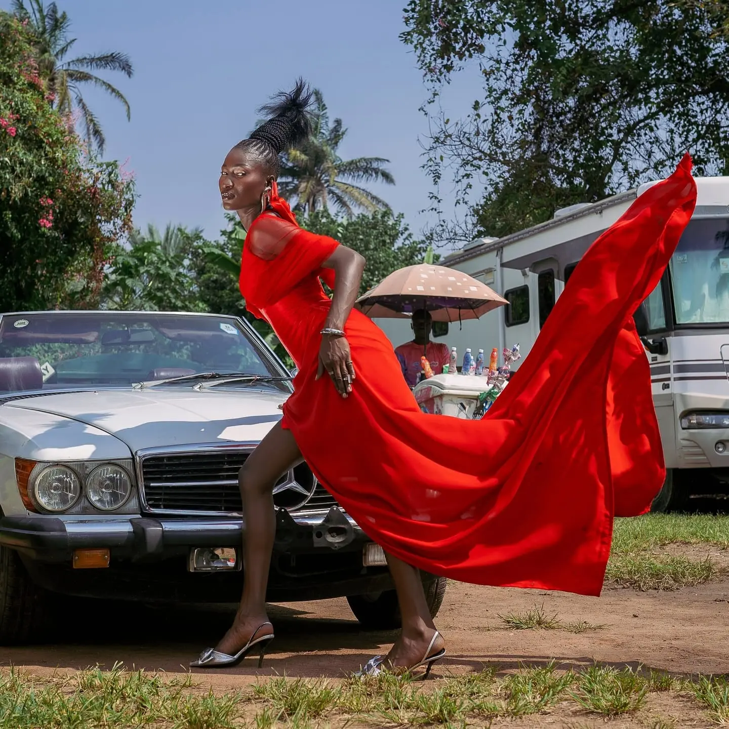 Model in a flowing red gown with sculpted braids strides past a vintage Mercedes convertible, fabric billowing behind her.