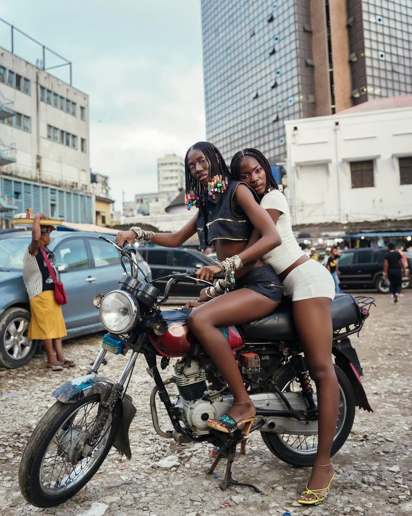 Two models with beaded braids share a motorcycle on a busy Lagos street, high-rises behind them.