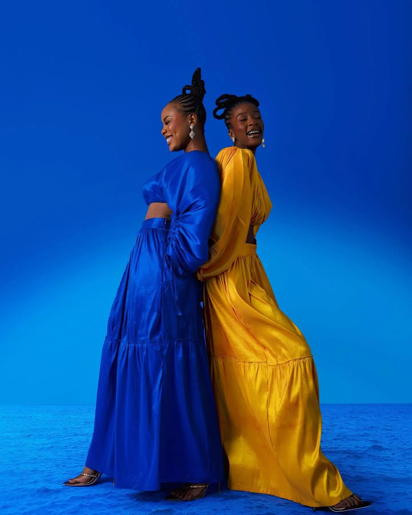 Two women in flowing cobalt-blue and golden-yellow gowns laugh back to back against a deep blue backdrop.