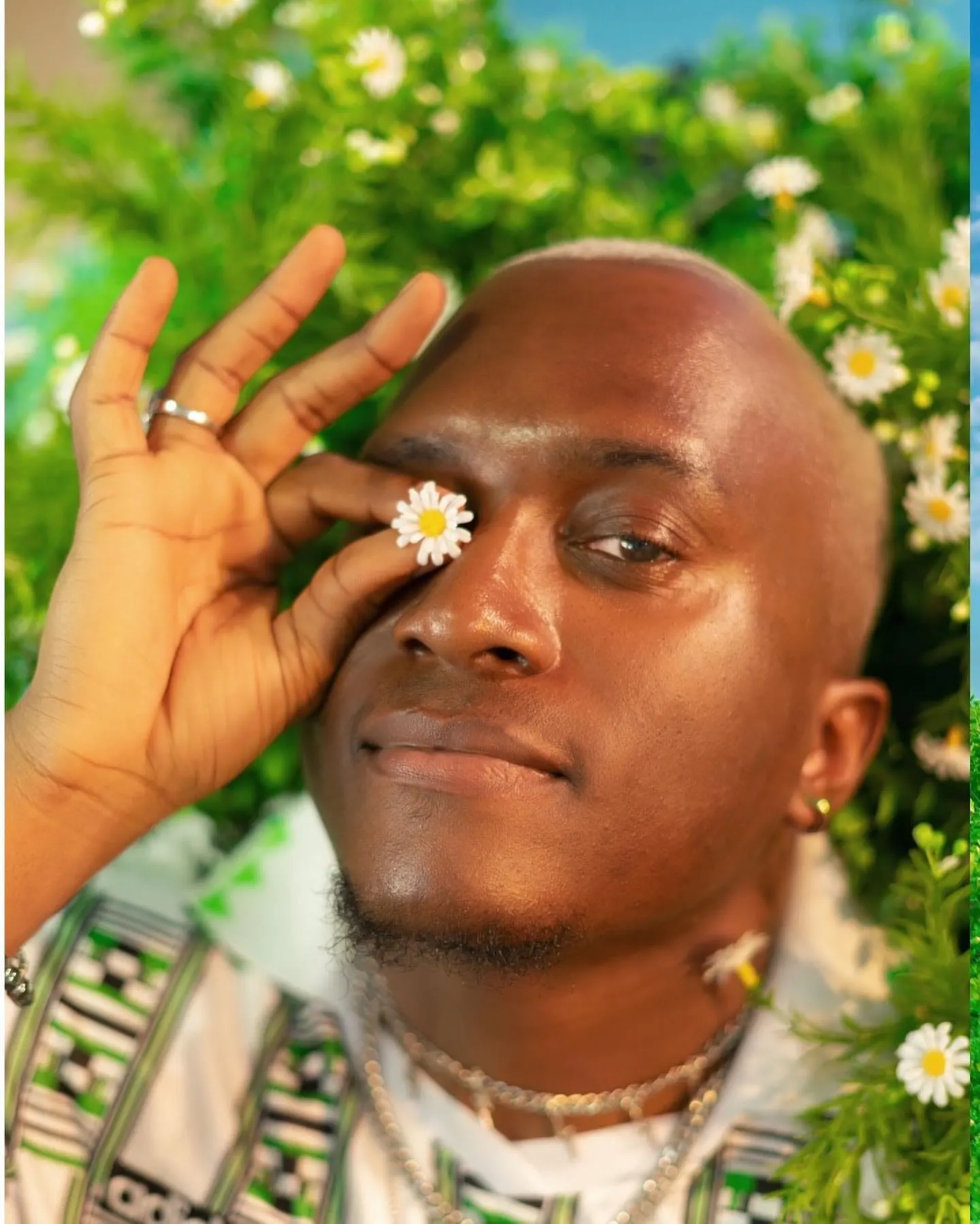 Man lying among daisies holds a single daisy over one eye, smiling softly.
