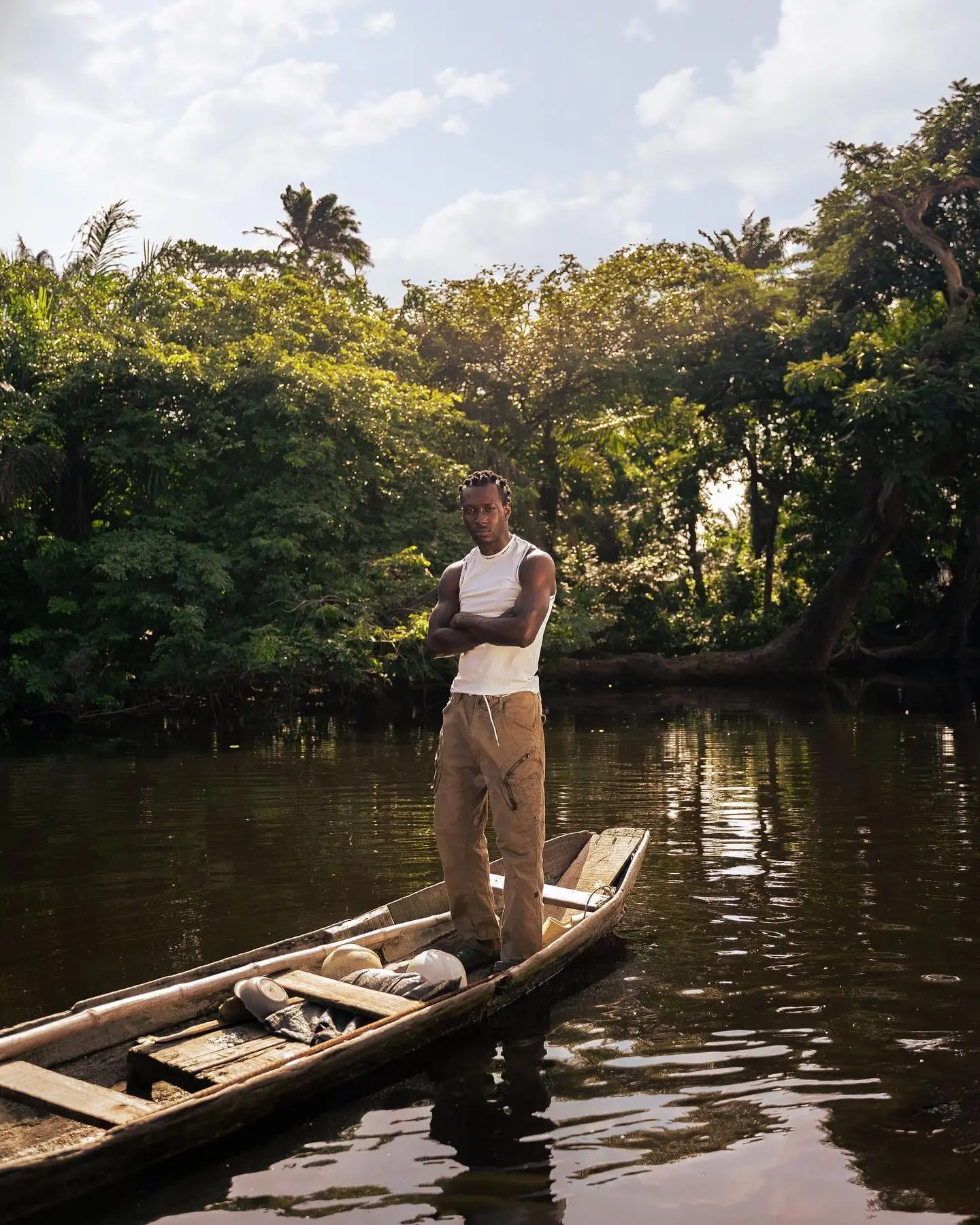 Man in a white vest stands with arms crossed in a wooden canoe on a calm forest-lined river.