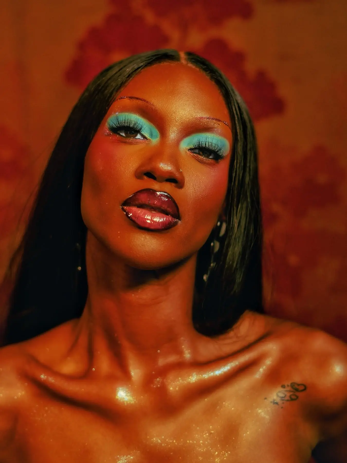 Close portrait of a woman with turquoise eyeshadow, glossy lips and glitter-dusted shoulders against warm red tones.