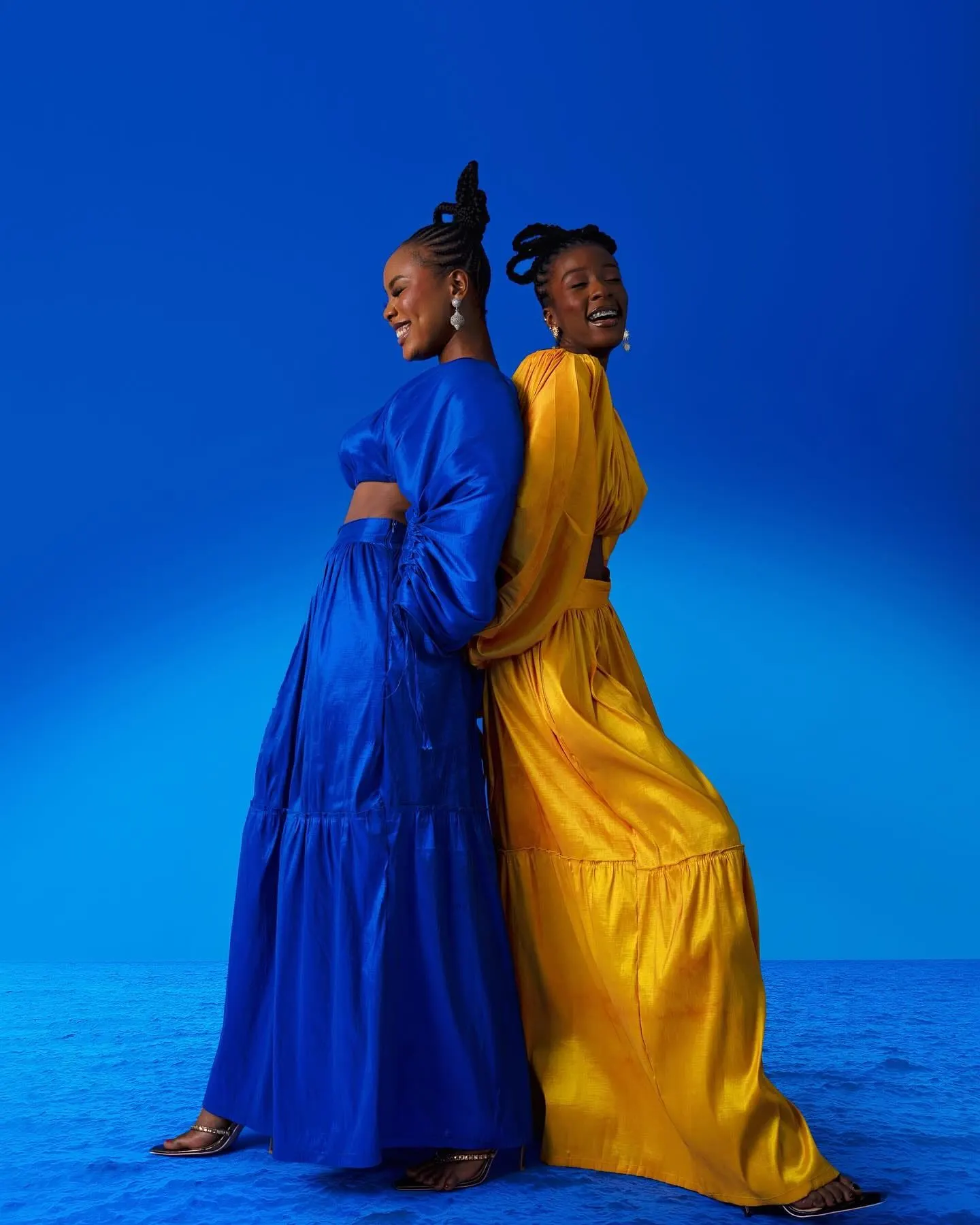 Two women in flowing cobalt-blue and golden-yellow gowns laugh back to back against a deep blue backdrop.
